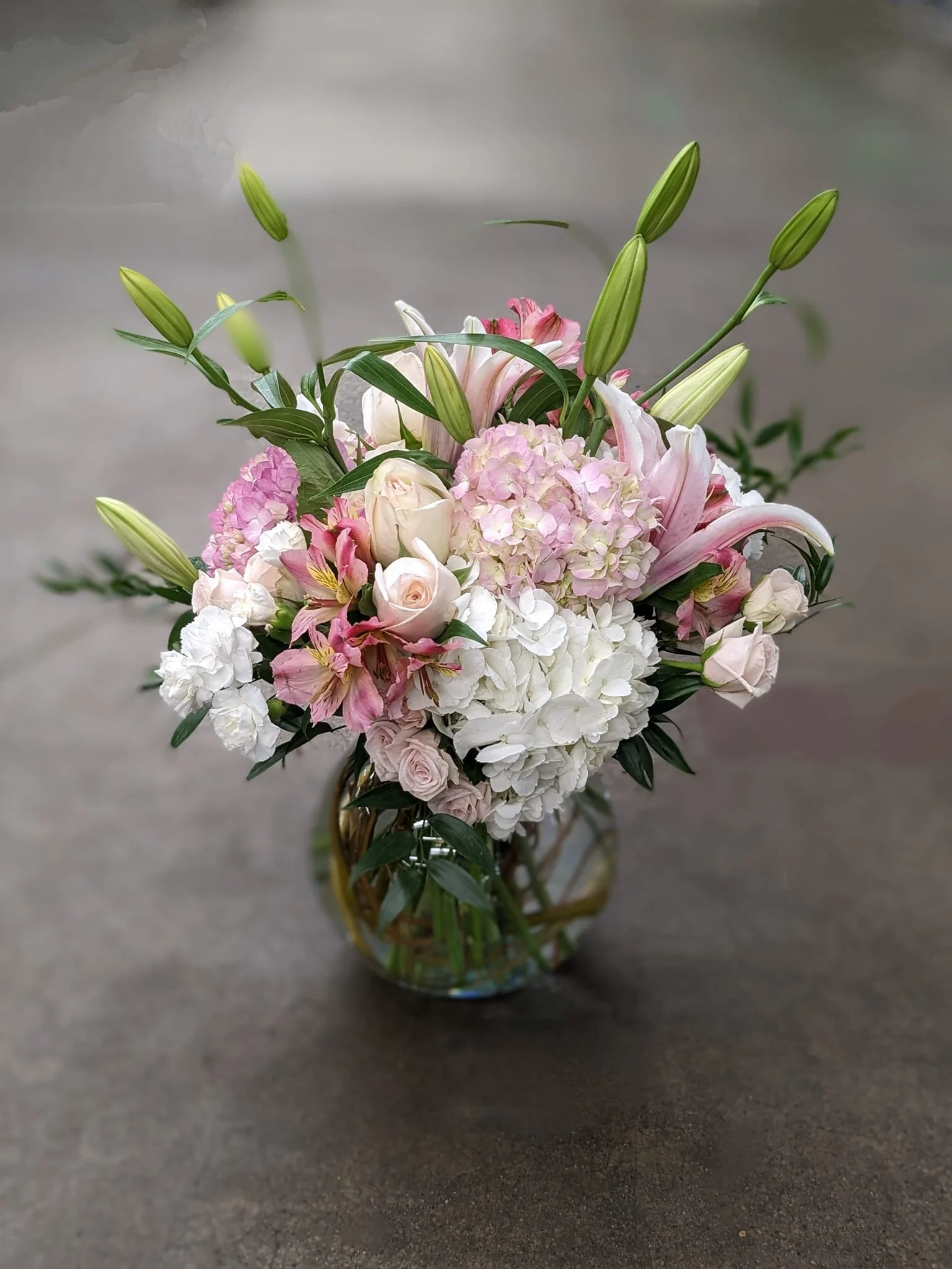 A bouquet of pink and white flowers, including lilies, roses, and hydrangeas, in a small glass vase on a dark surface.