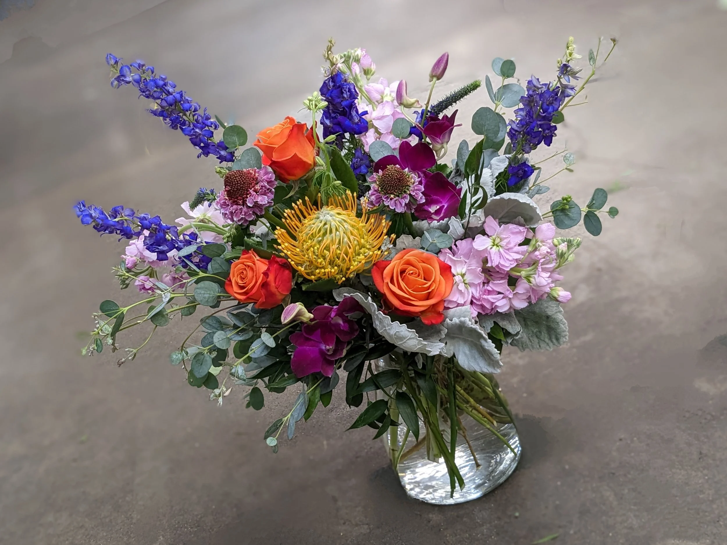 A colorful bouquet of flowers in a glass vase on a dark surface, featuring orange roses, blue delphiniums, pink astilbes, purple calla lilies, and green eucalyptus leaves.