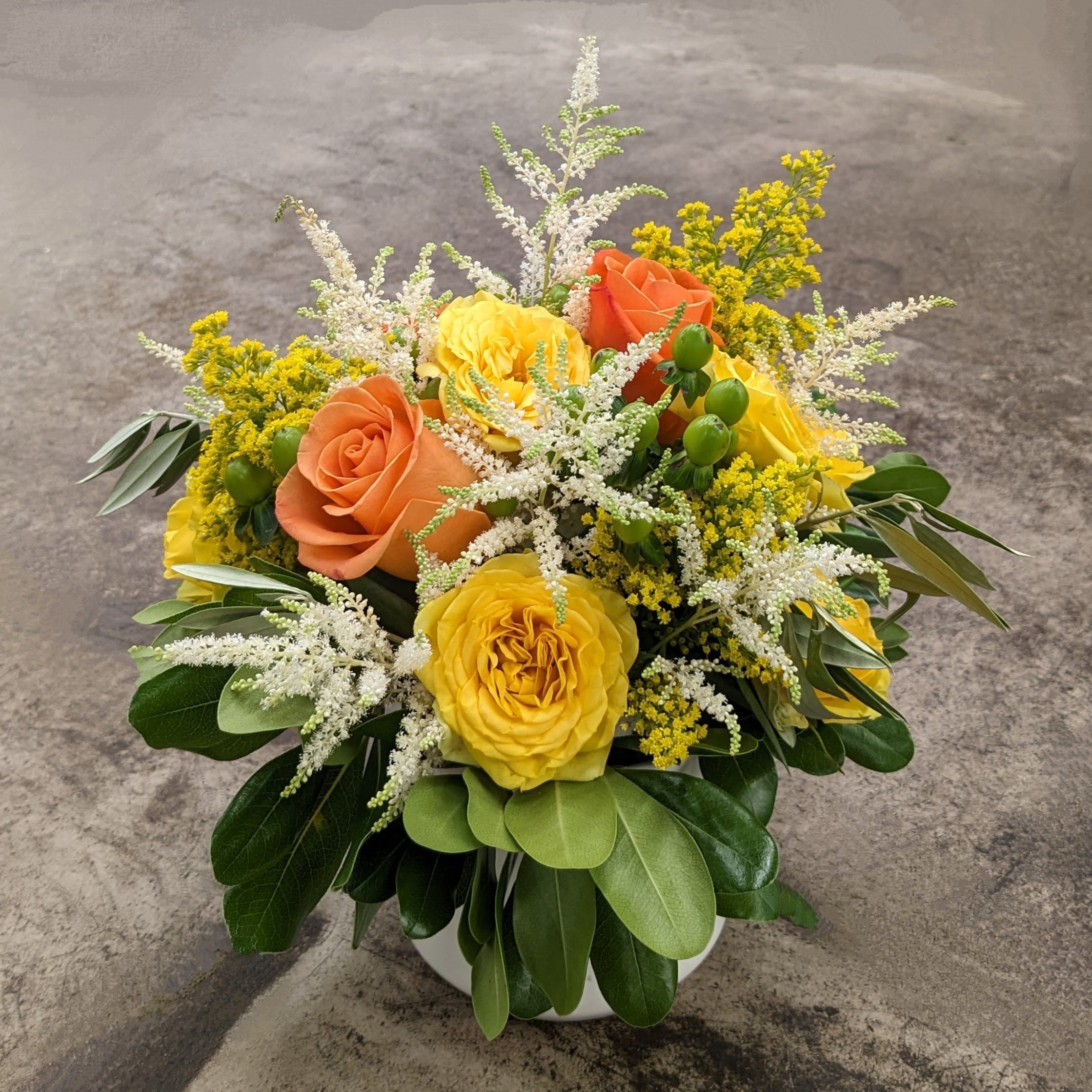 A bouquet of yellow, orange, and white flowers with green leaves in a white vase on a gray surface.