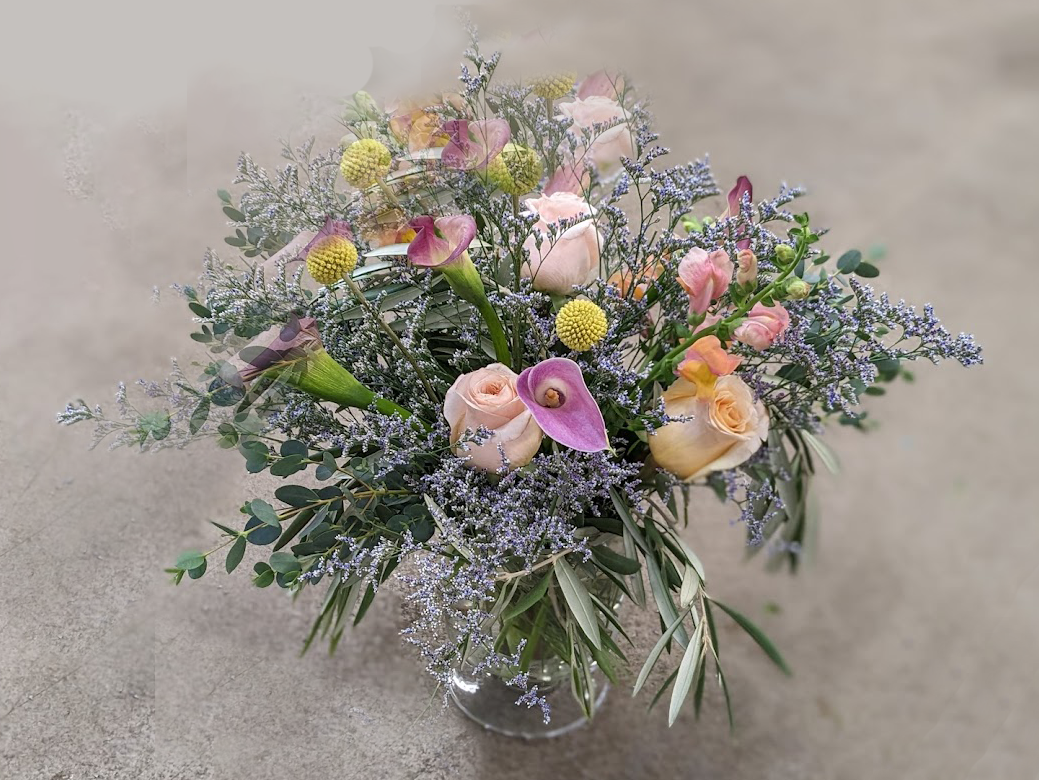 A floral bouquet in a glass vase containing pink calla lilies, pale peach roses, pink and peach tulips, and small yellow billy buttons, with purple and green foliage.