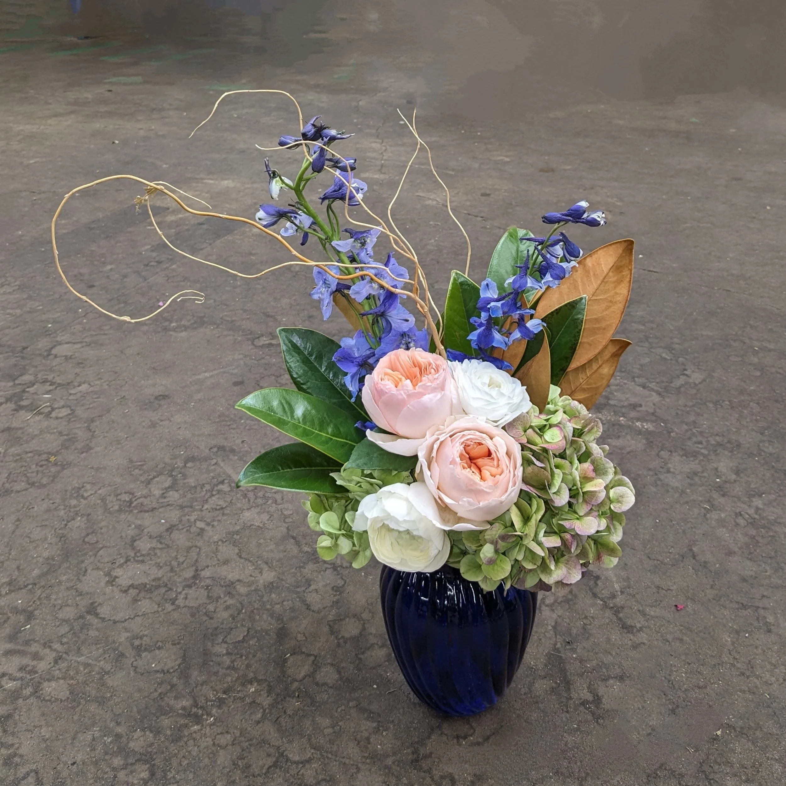 A bouquet of flowers in a blue vase with purple, white, and pink flowers, green leaves, and dried curly tendrils on a concrete surface.