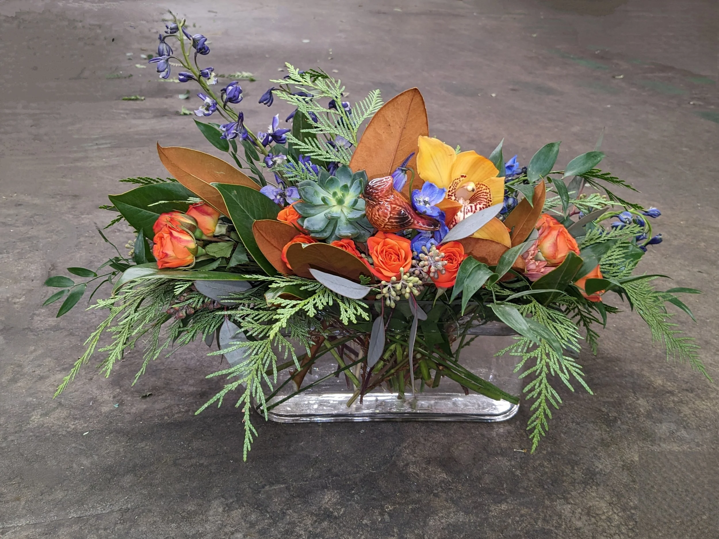 A floral arrangement in a clear glass rectangular vase featuring purple flowers, orange roses, yellow orchid, green succulent, orange bird figurine, and green leaves and ferns on a dark surface.