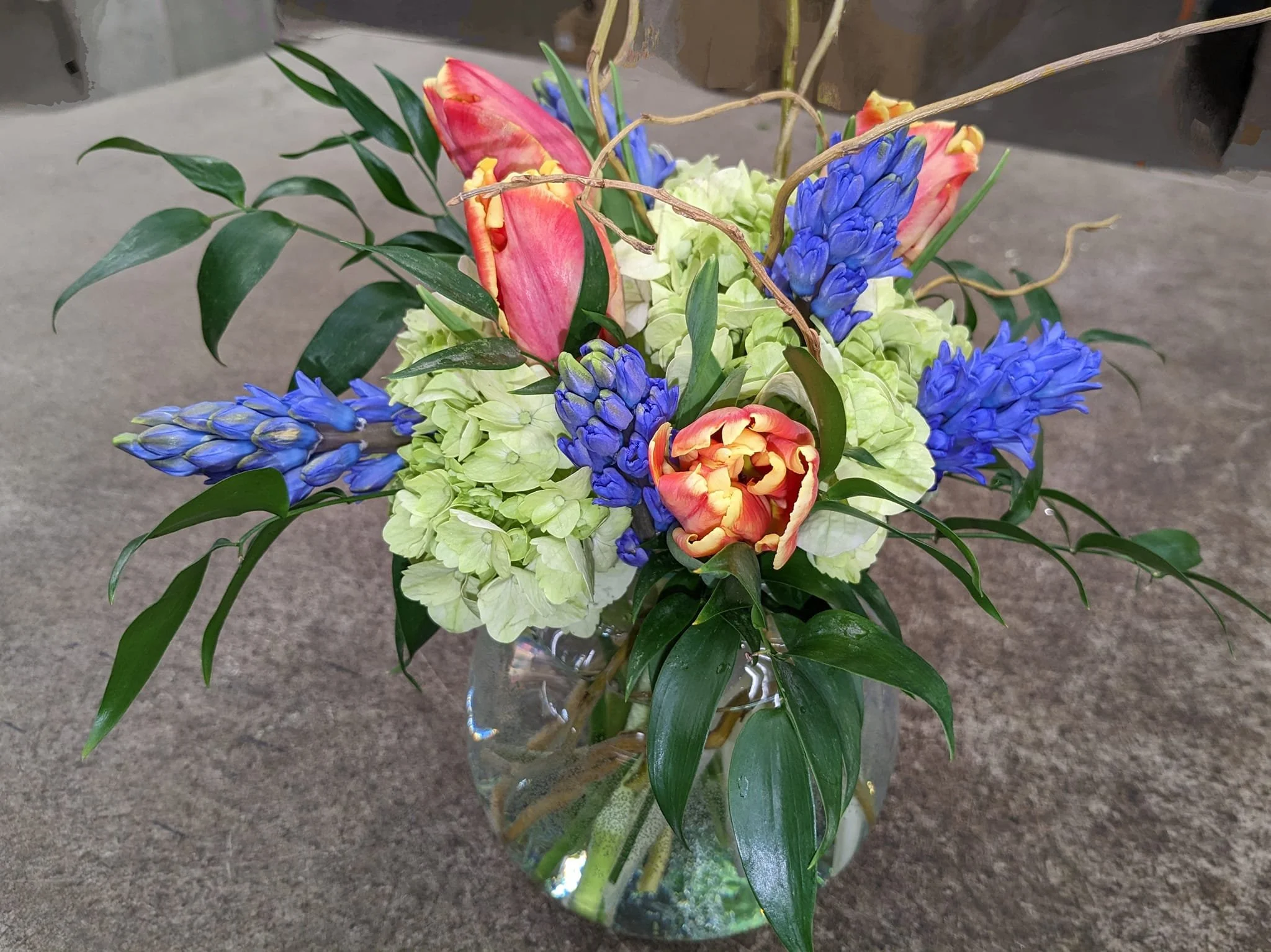 A floral arrangement in a glass vase containing pink tulips, blue hyacinths, green hydrangeas, and various green leaves and twigs.