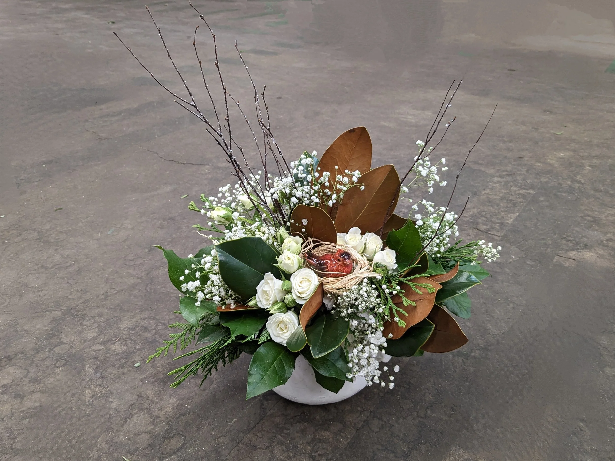 A floral arrangement in a white pot, featuring white roses, baby's breath, green leaves, magnolia leaves, and sticks, with a small decorative frog figurine nestled among the flowers.