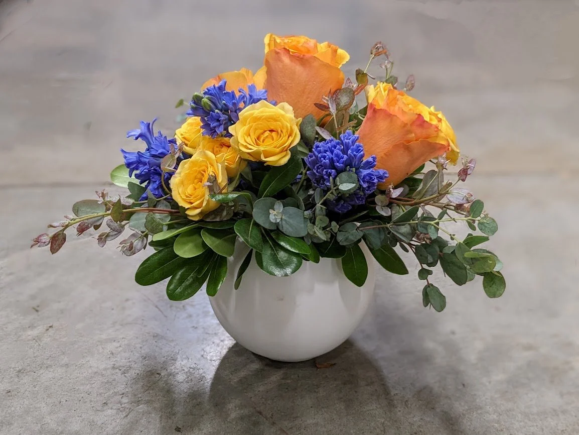 A small white vase containing a bouquet of yellow roses, orange tulips, purple hyacinths, and green foliage.
