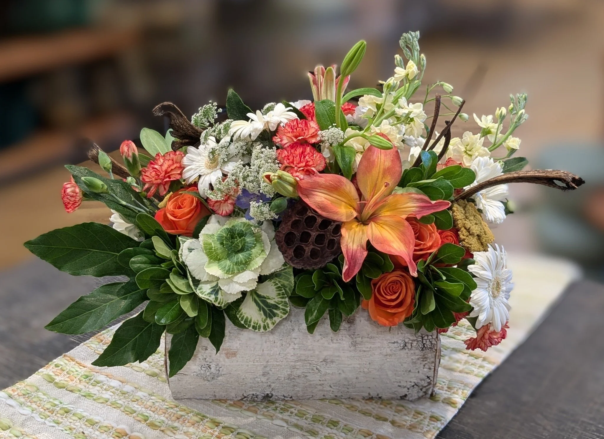 A colorful bouquet of fresh flowers, including orange roses, pink and yellow lilies, white daisies, and other mixed blossoms, arranged with green foliage and placed on a rustic wooden surface with a patterned fabric underneath.