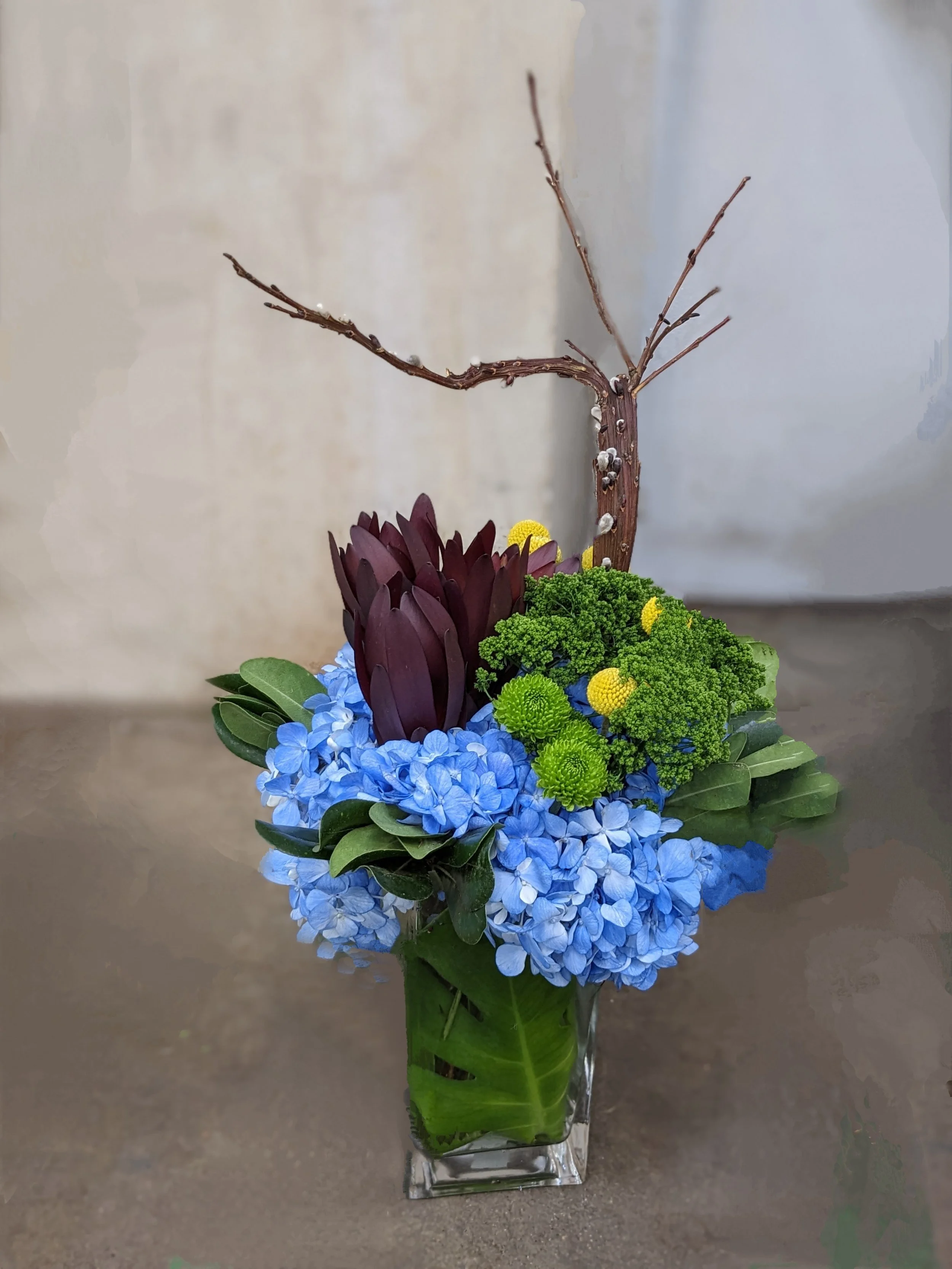 A floral arrangement in a glass vase with blue hydrangeas, green chrysanthemums, purple succulents, and a large leaf wrapped at the bottom, with a bare tree branch extending upward.