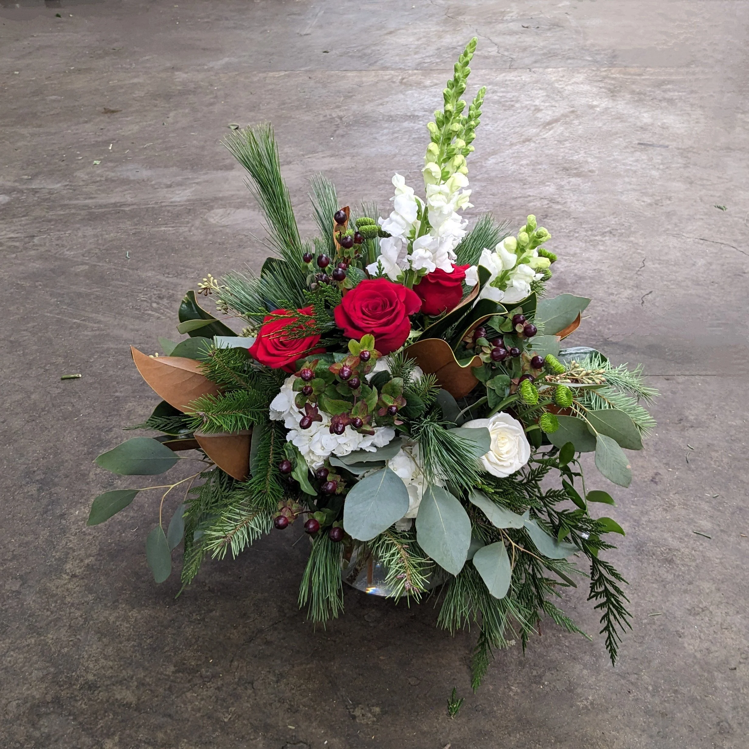 A floral arrangement with red roses, white snapdragons, greenery, and other small flowers on a concrete floor.