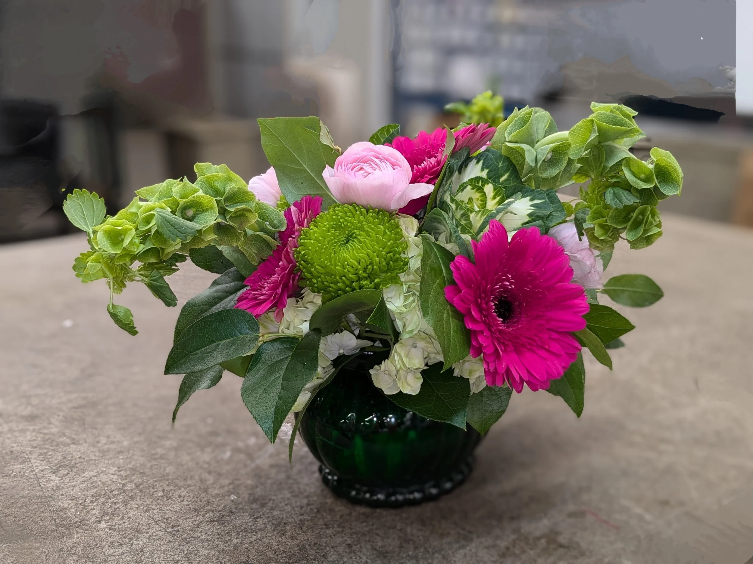 A bouquet of pink, white, and green flowers in a dark green glass vase on a gray surface.