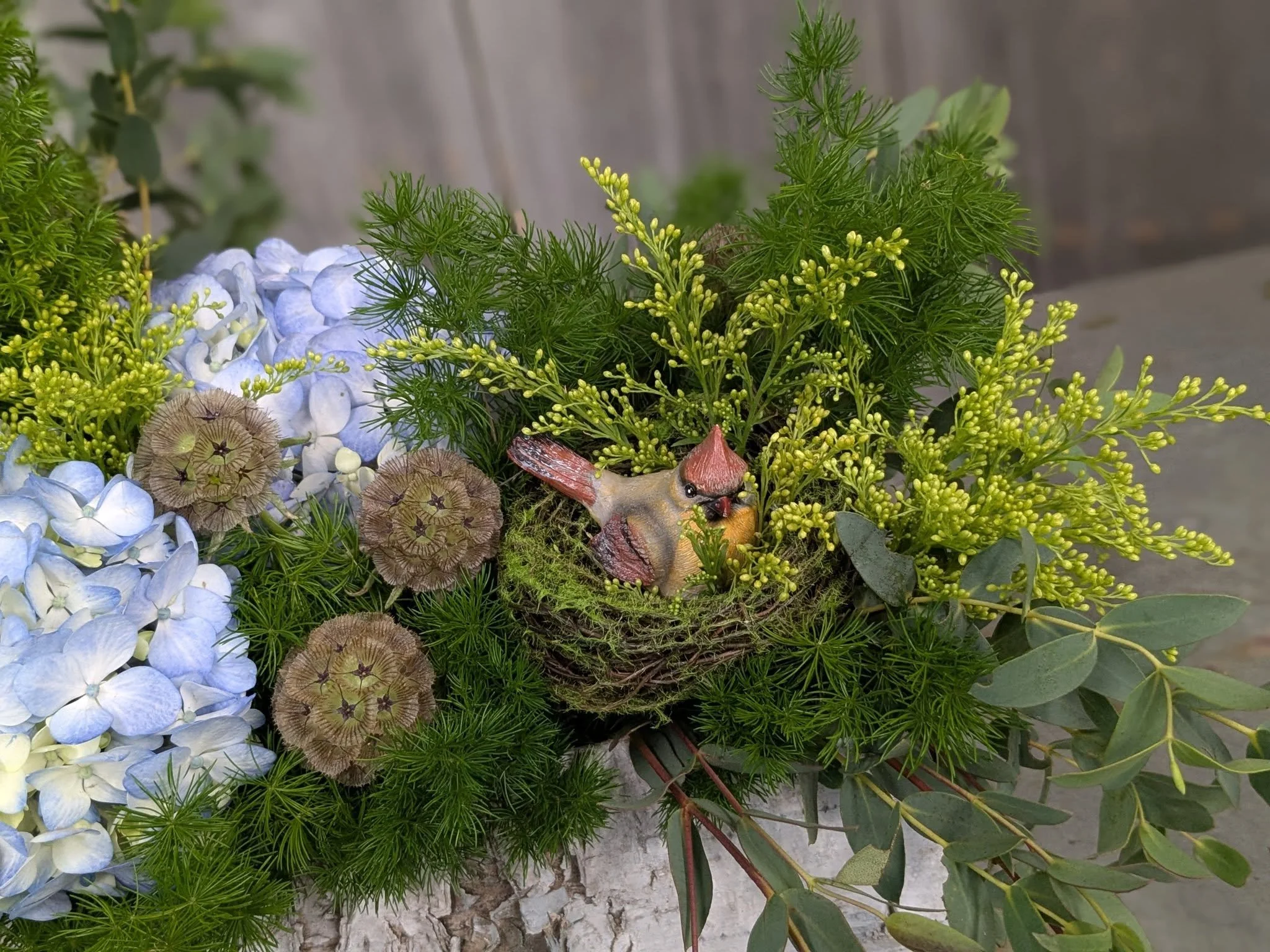 A floral arrangement with blue hydrangeas, green foliage, and a small decorative bird among the flowers, set on a concrete surface.