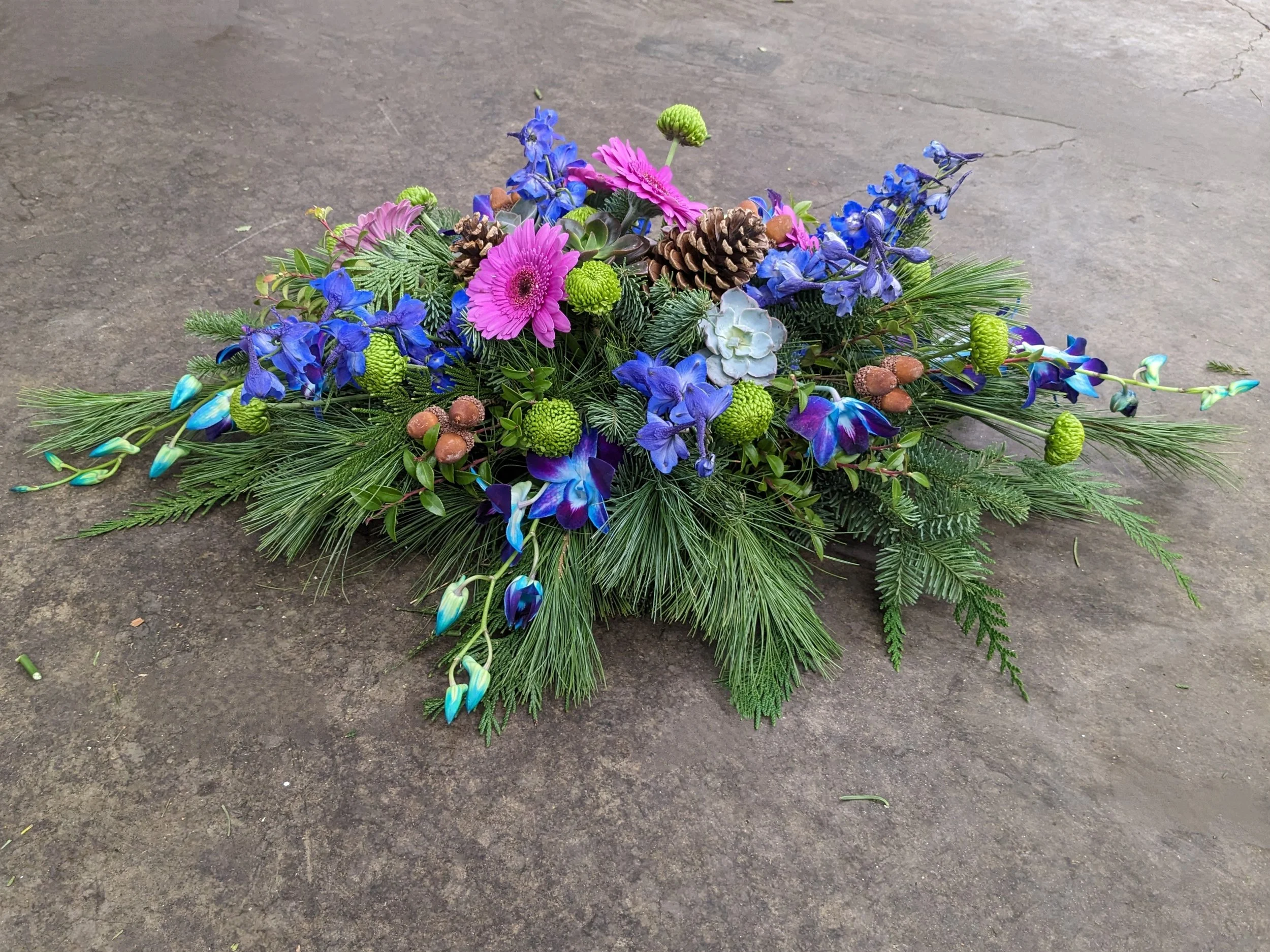 A floral arrangement on a concrete surface featuring pink, purple, and blue flowers, pinecones, green ornamental balls, and greenery.