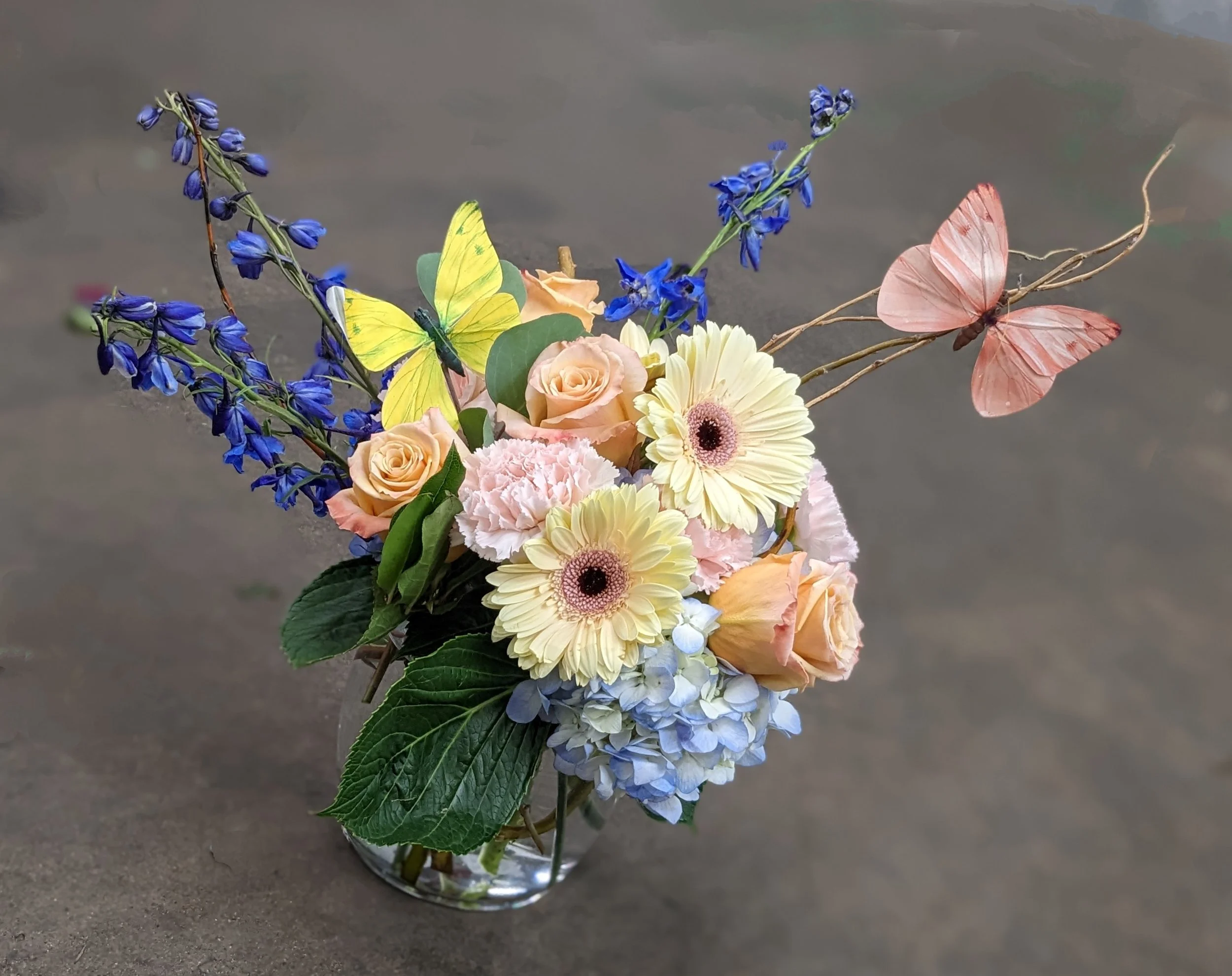 A bouquet of flowers including blue delphiniums, yellow daisies, peach roses, pink carnations, and two pink butterflies with extended wings, set against a gray background.