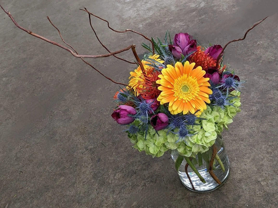 Colorful flower arrangement in a glass jar containing yellow, purple, and green flowers along with decorative branches.