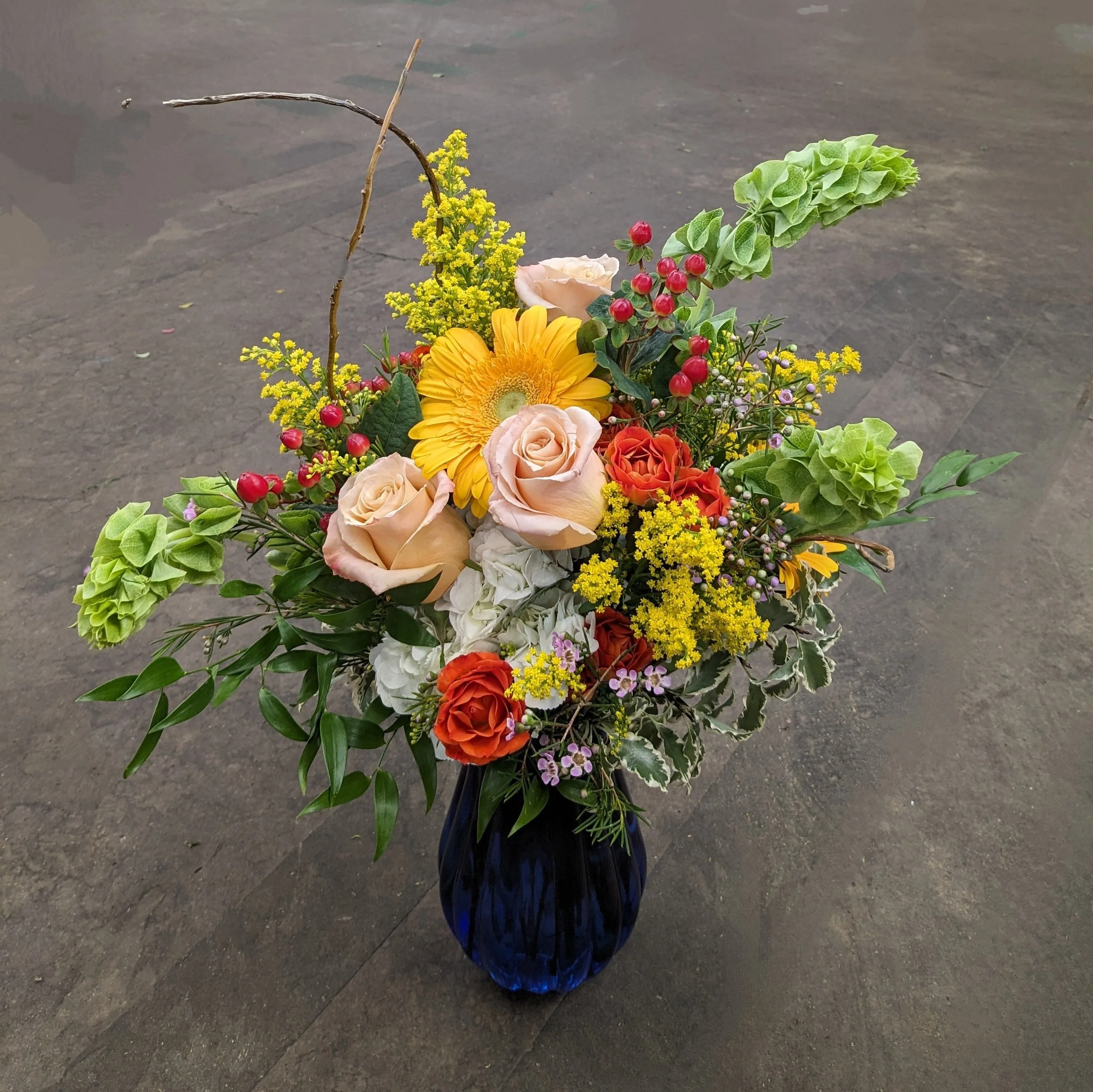 A floral arrangement in a tall, blue glass vase, containing yellow, pink, and orange flowers, green leaves, red berries, and thin branches over a dark pavement background.