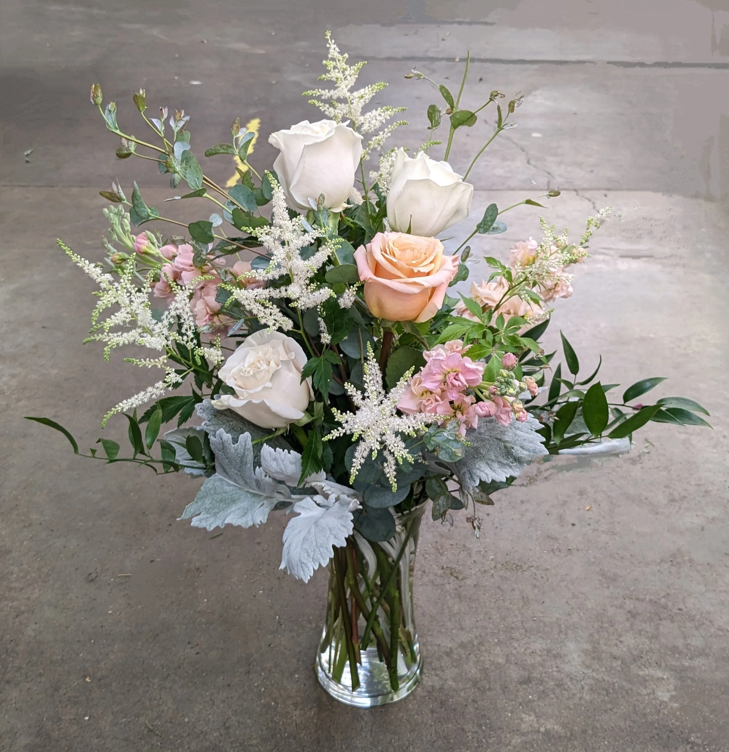 A bouquet of white and pale pink roses, white daisies, green leaves, and other small white flowers arranged in a clear glass vase, placed on a concrete floor.