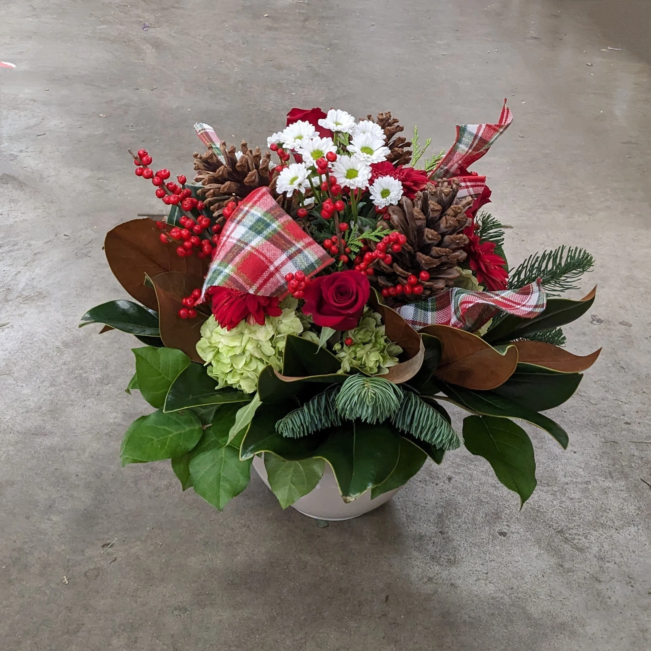 A decorative Christmas flower arrangement with white daisies, red berries, pinecones, red roses, greenery, and plaid ribbons in a container, placed on a concrete floor.