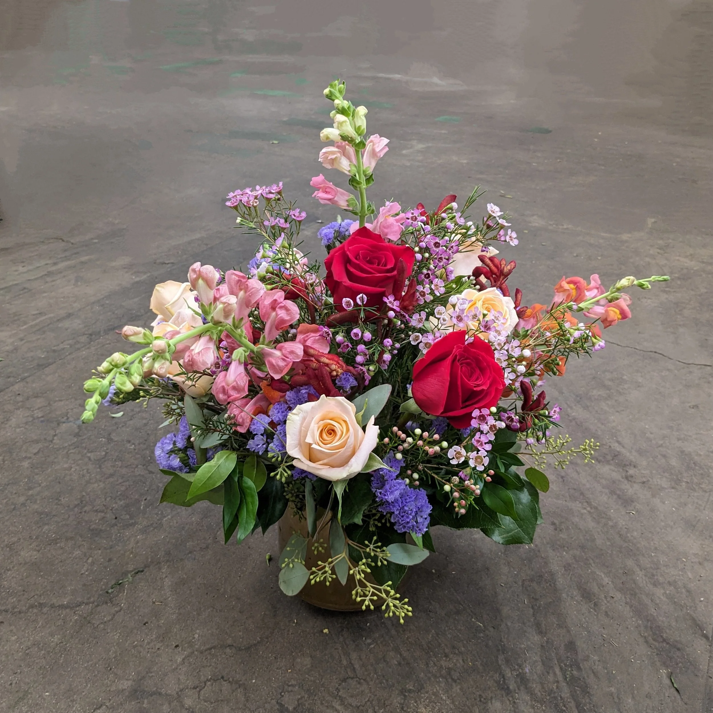 A floral arrangement with various flowers including pink, red, purple, cream, and peach roses, along with other small colorful blossoms, arranged in a bowl on a rough concrete surface.