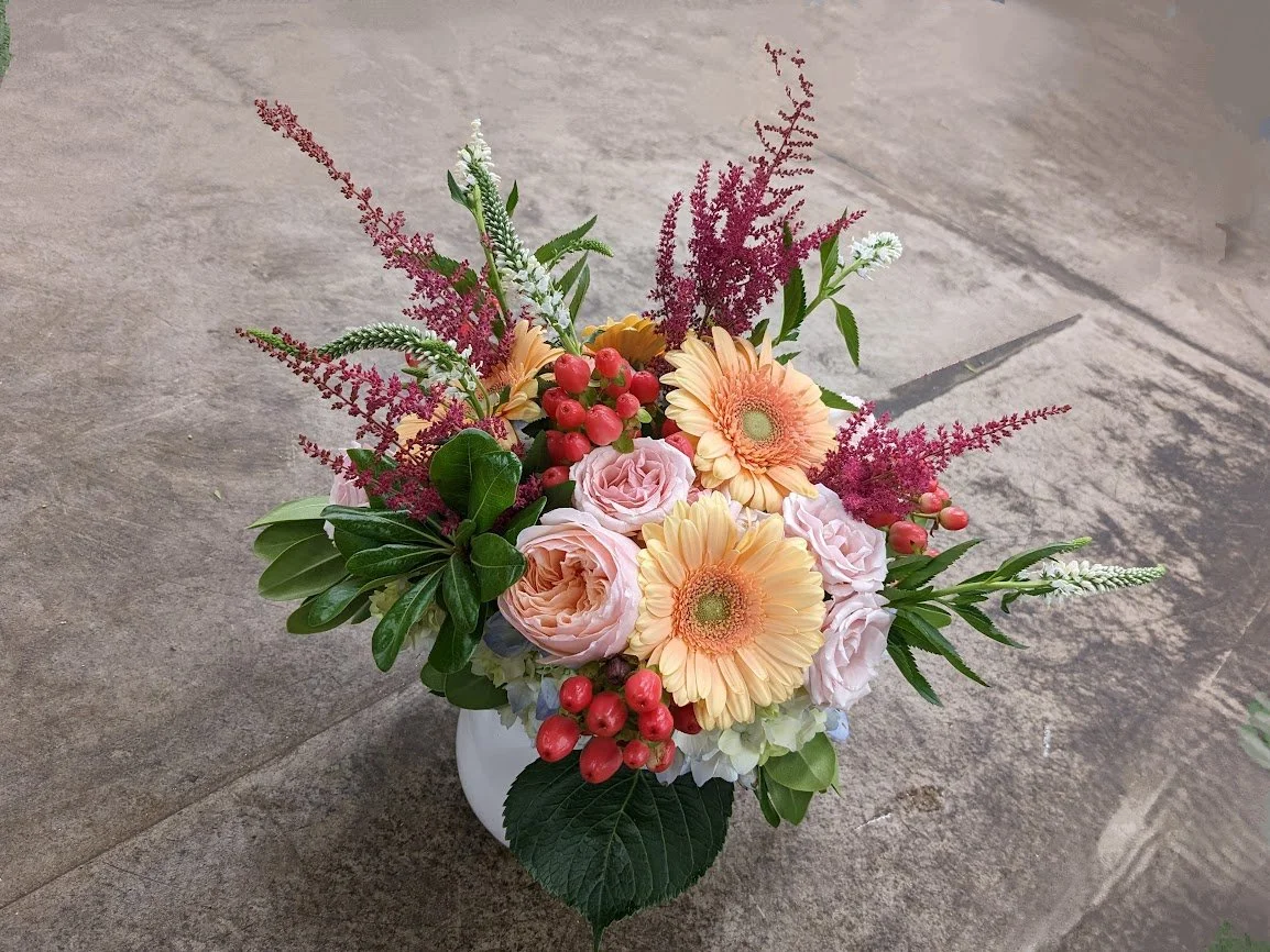 A bouquet of pink roses, orange gerbera daisies, red berries, green leaves, and purple, white, and pink fillers in a white vase on a stone surface.