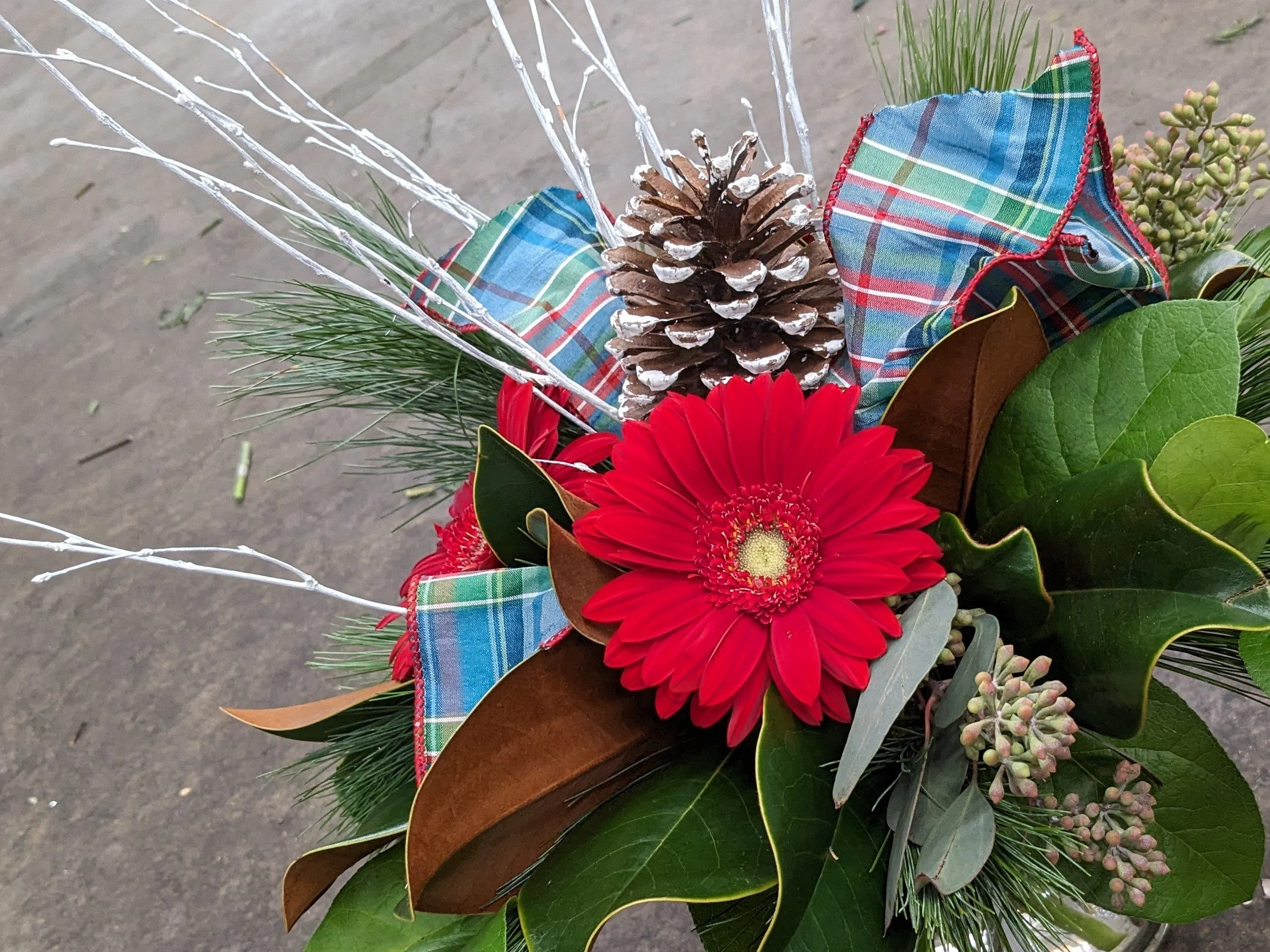 A floral arrangement with red flowers, pinecone, green pine needles, and plaid fabric accents, set against a gray outdoor surface.