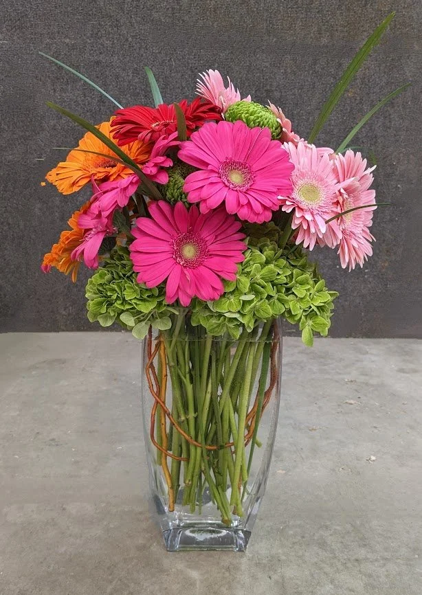 A clear glass vase filled with colorful flowers, including pink, orange, and light purple gerbera daisies, and green hydrangeas, set against a gray background.