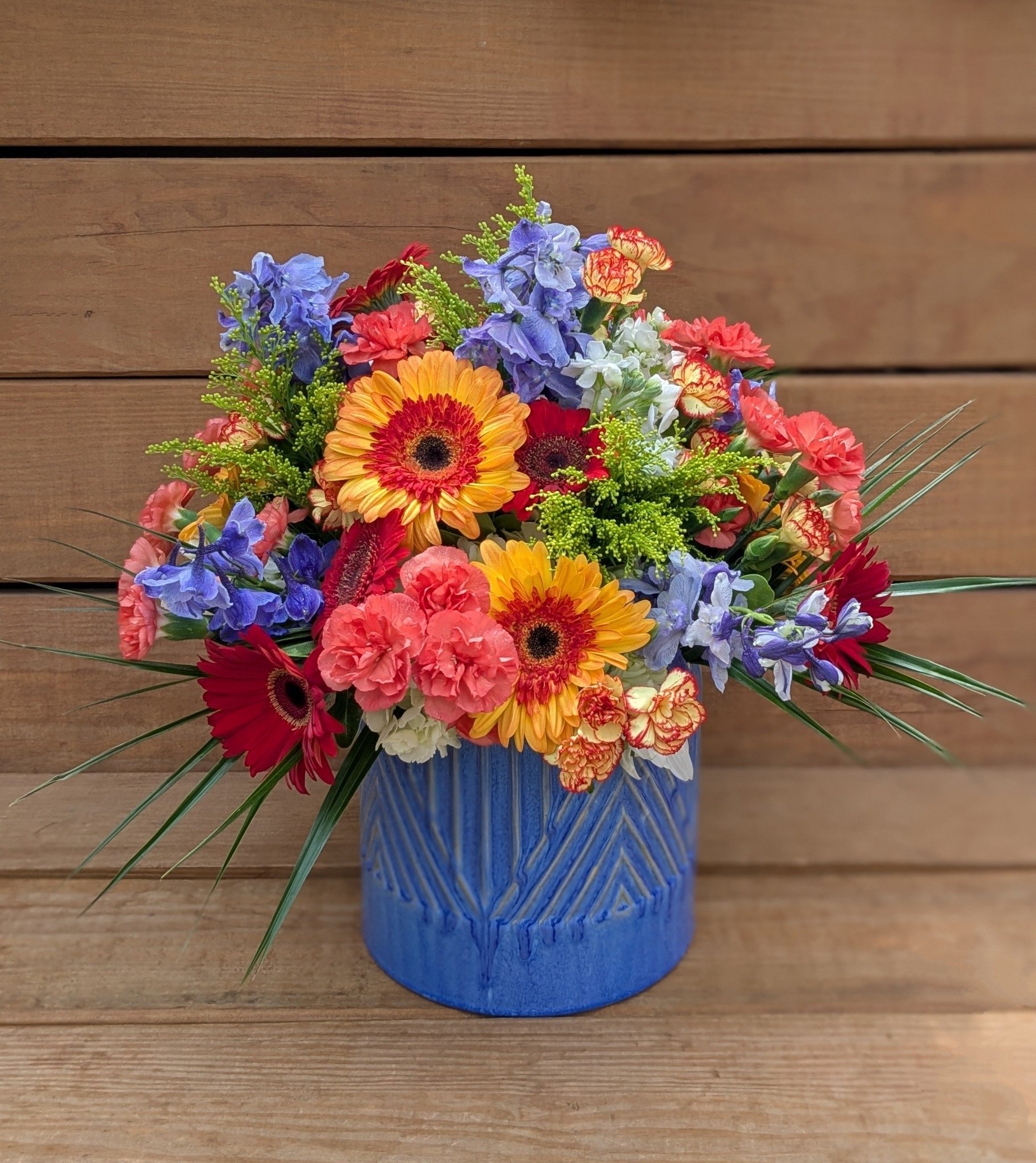 A vibrant bouquet of mixed flowers including orange gerbera daisies, red carnations, purple delphiniums, and pink and yellow carnations in a blue ceramic vase on a wooden surface.
