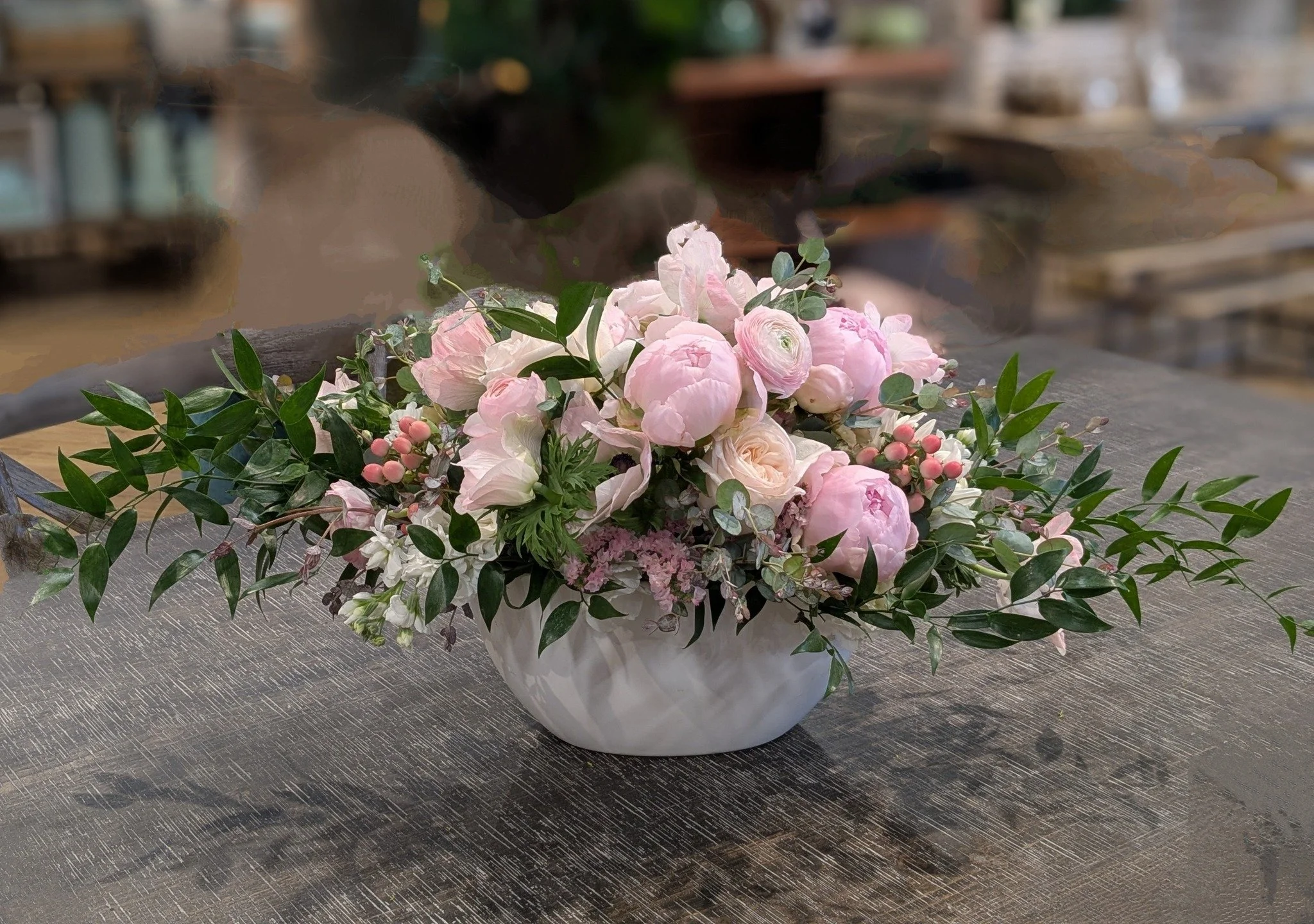 A floral arrangement in a white ceramic vase containing pink peonies, white roses, pink berries, and green foliage, placed on a textured dark surface.
