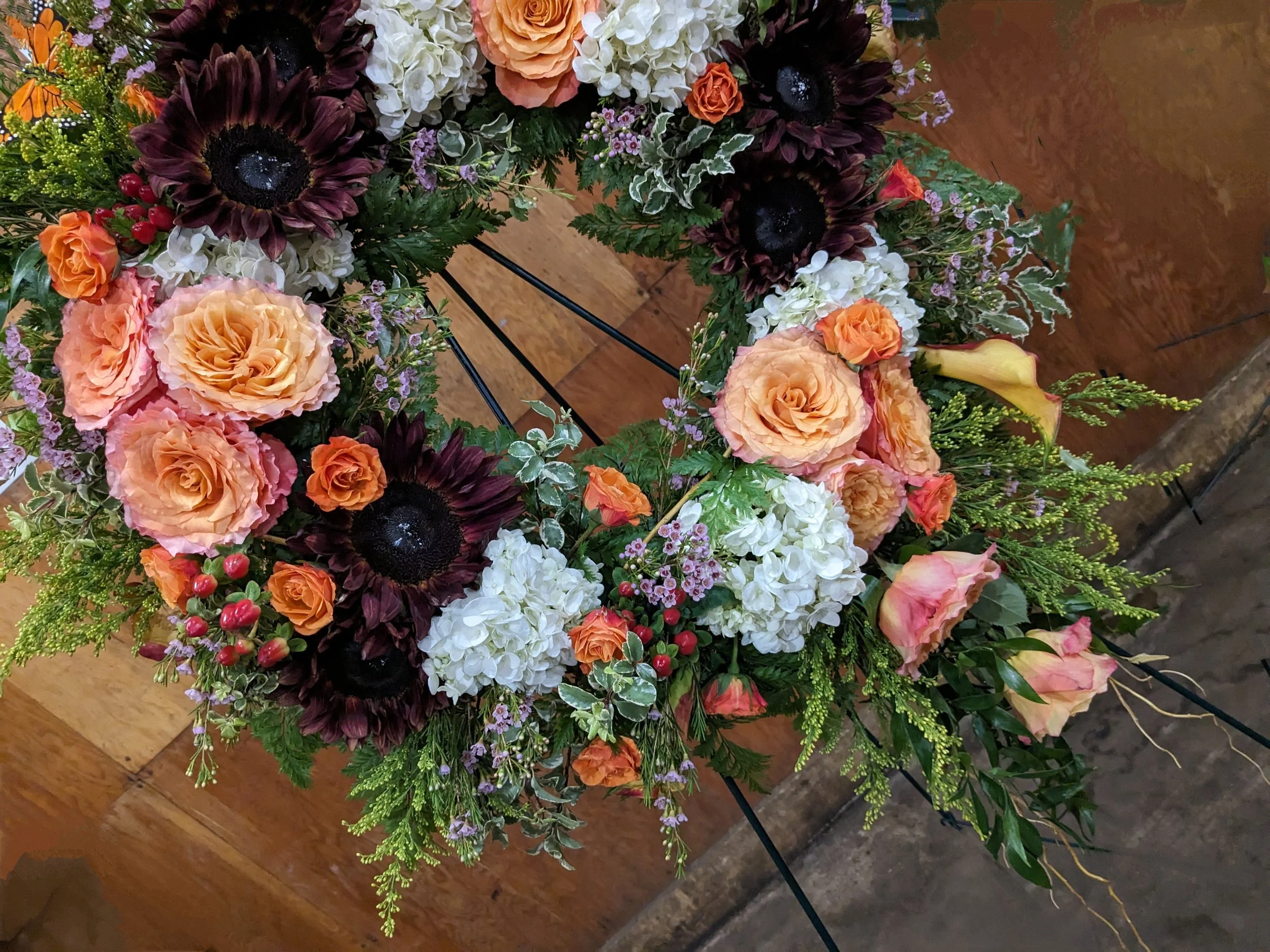 A floral arrangement with various flowers, including large peach roses, dark burgundy sunflowers, white hydrangeas, small purple flowers, red berries, and assorted green foliage, placed on a wooden surface.