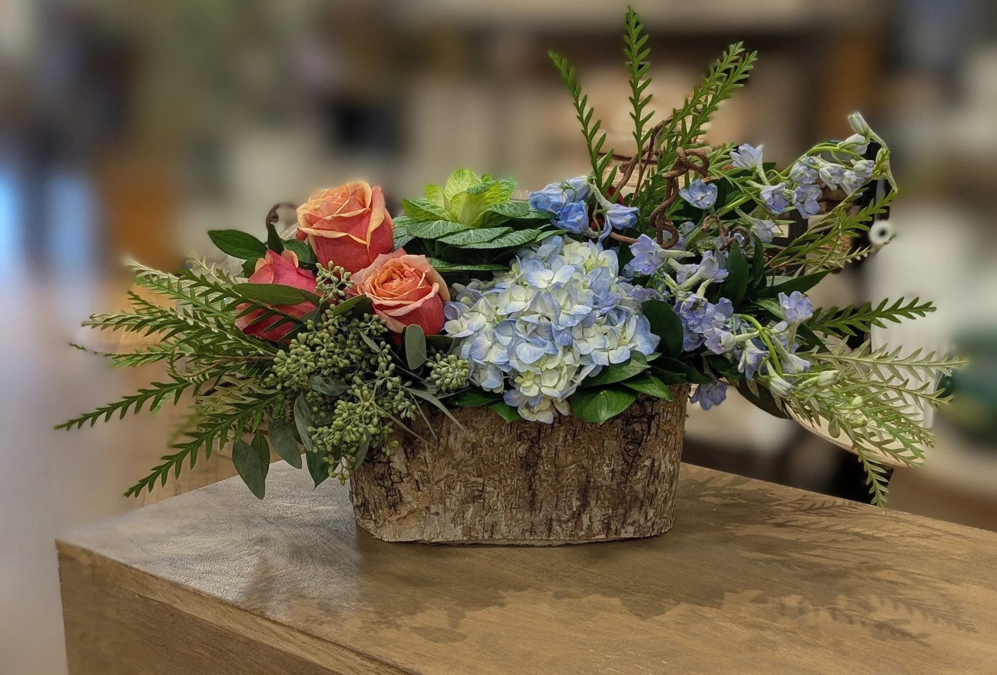 A floral arrangement with pink roses, blue hydrangeas, and green foliage in a rustic wood container on a wooden table.
