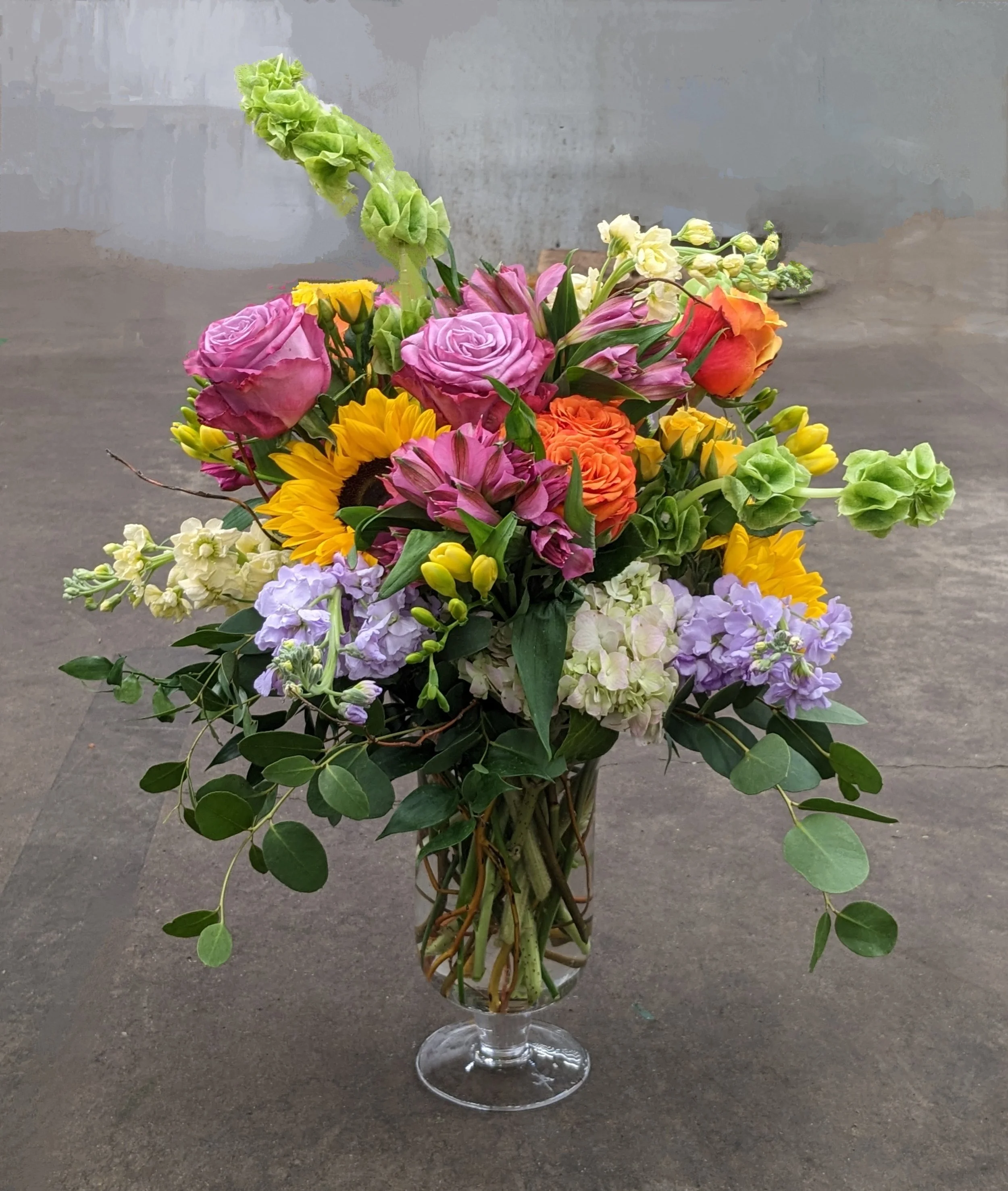 A bouquet of colorful flowers, including pink roses, sunflowers, orange roses, purple and white hydrangeas, and white stock, arranged in a large glass vase on a gray surface.