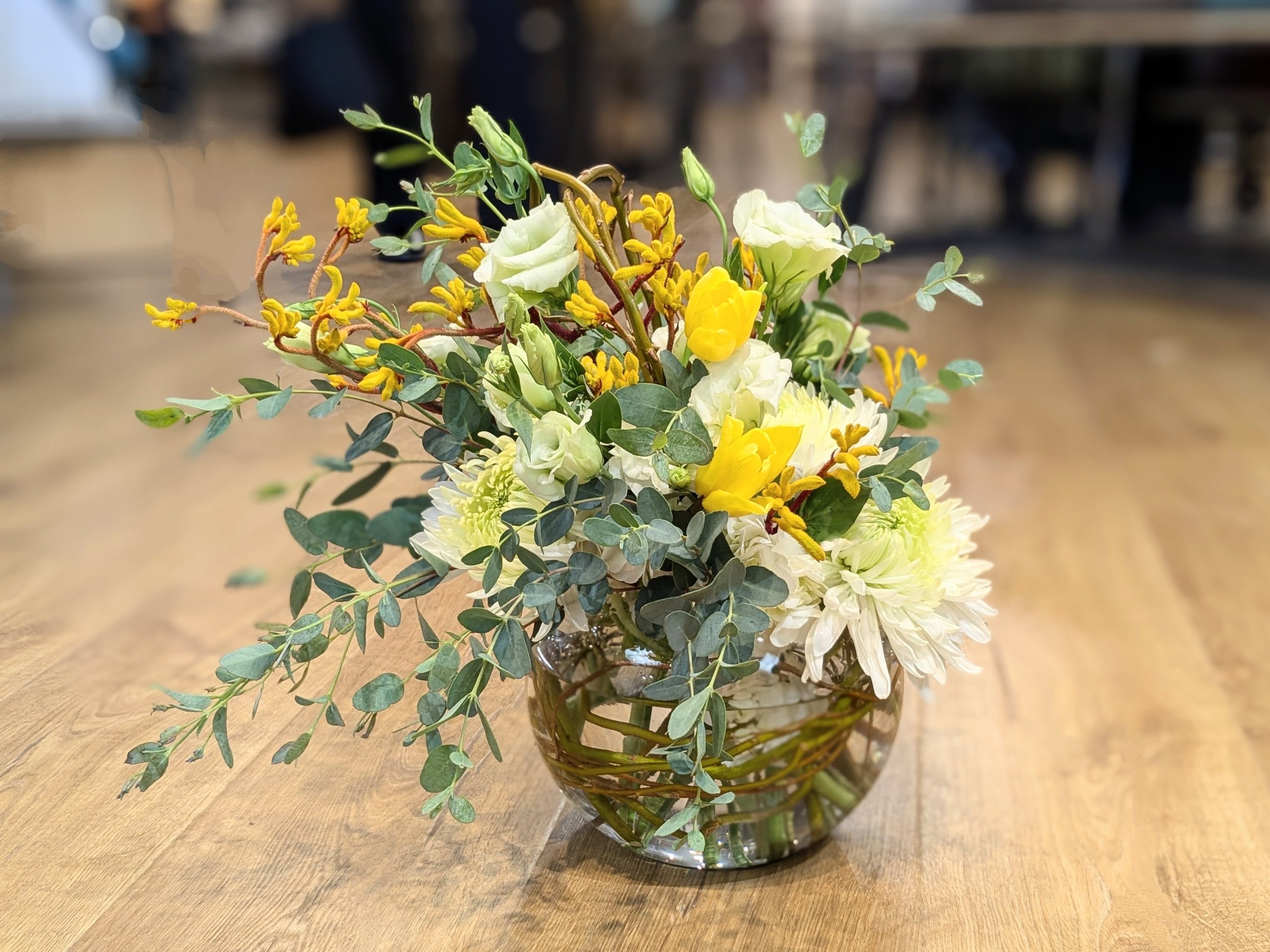 A glass vase filled with white and yellow flowers, green leaves, and yellow accents, placed on a wooden surface.