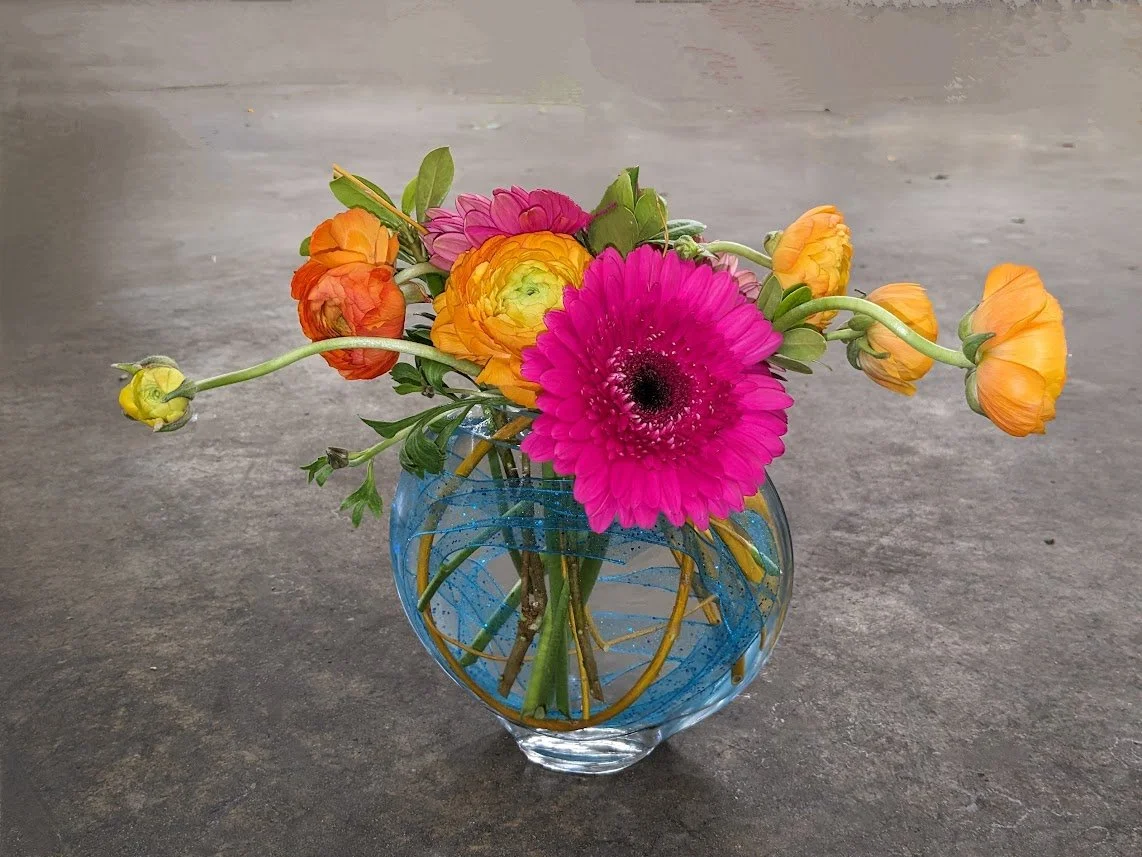 Colorful floral bouquet in a glass vase with pink, orange, and yellow flowers on a gray surface.