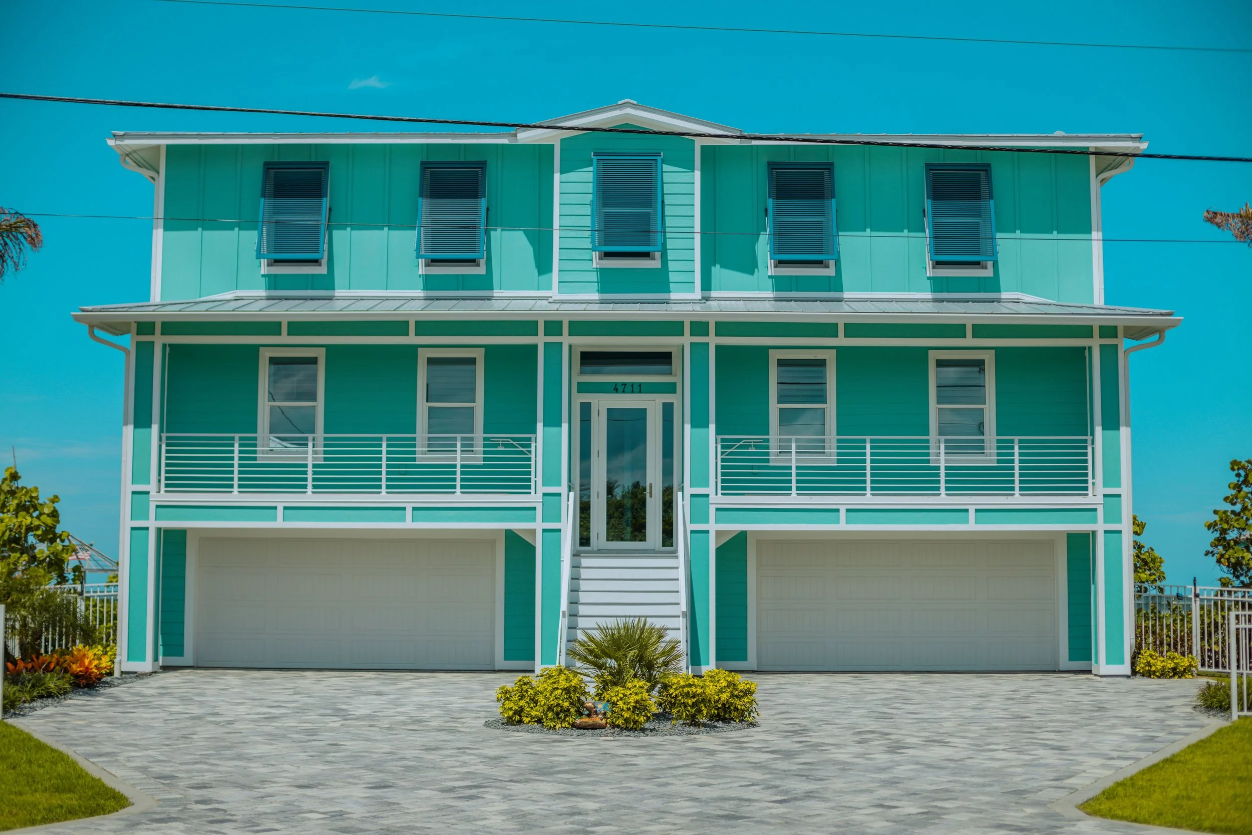 Front view of a turquoise-colored, two-story house with a white railing and a garage door. The house has four windows with black shutters on the upper floor and two on the lower floor. There are small landscaped bushes and greenery in front of the house, and the driveway is paved with stone tiles.