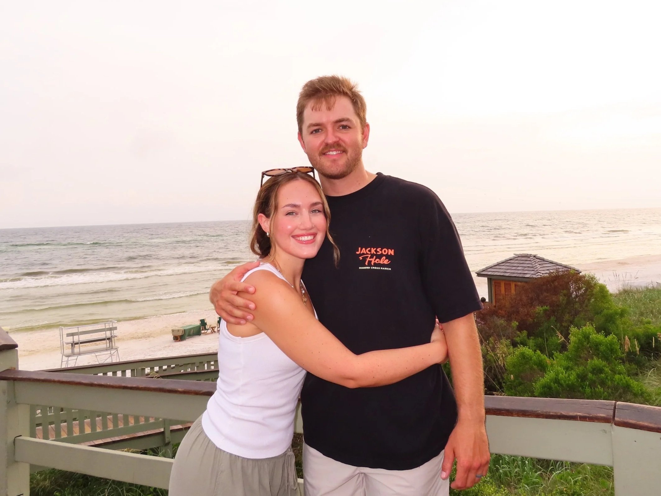 A smiling couple hugging by the beach with ocean waves, a small structure, and a sandy shoreline in the background.