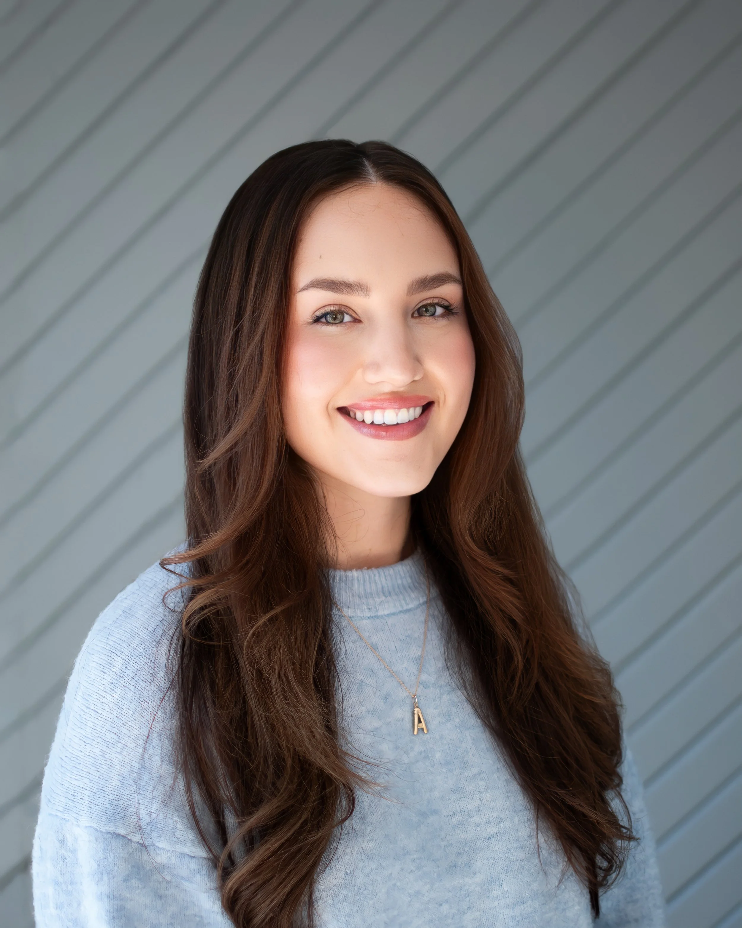 A young woman with long brown hair and light-colored eyes is smiling in front of a gray, slatted wall. She is wearing a light gray sweater and a gold necklace with a pendant shaped like the letter 'A'.