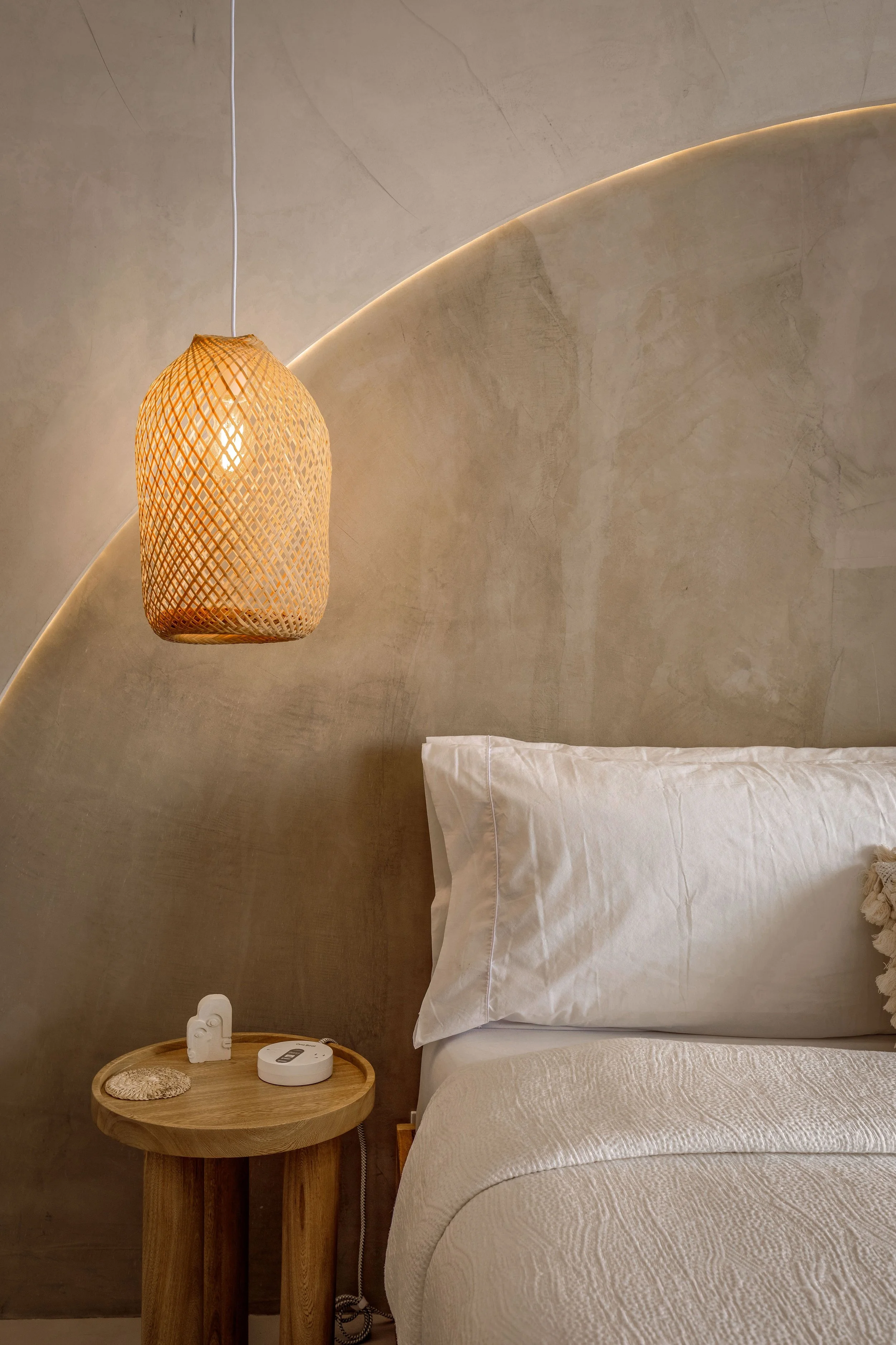 Close-up of a bedroom with a white bed, beige textured wall, hanging wicker pendant light, and a small round wooden bedside table with a clock, a small decorative item, and a switch.