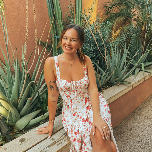 A woman with brown hair in a floral dress smiling and sitting on a wooden bench next to large green agave plants.