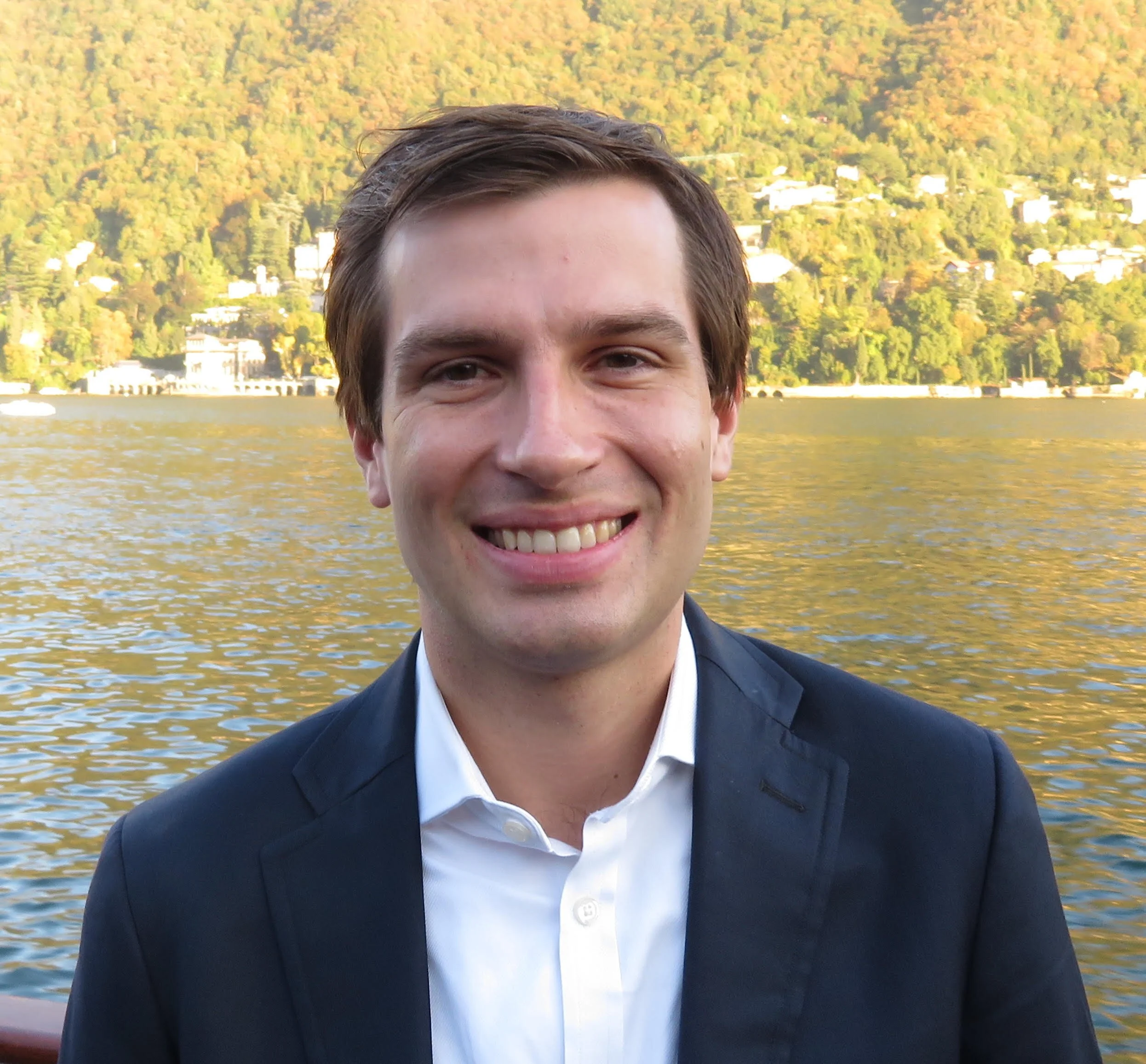 Close-up of a smiling young man in a dark suit and white shirt outdoors near water with a wooded hillside in the background.