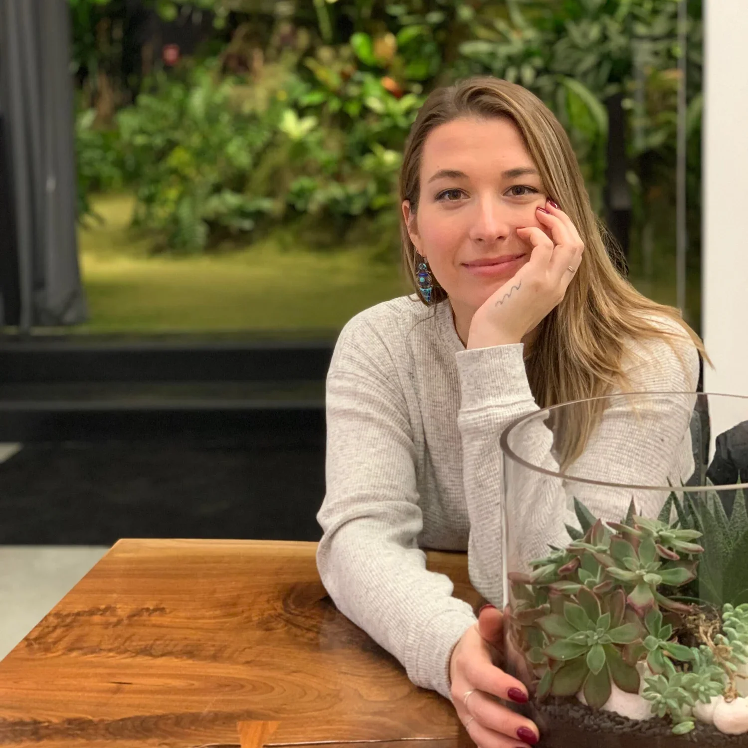 A woman with long brown hair, wearing earrings and a light long-sleeve shirt, sitting at a wooden table with a potted succulent and a glass container, smiling at the camera in front of a large window revealing greenery outside.