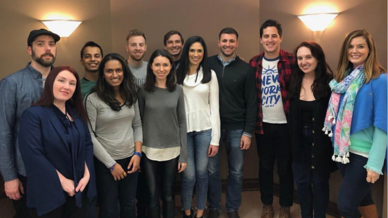 Group of fifteen young adults posing for a photo indoors, smiling at the camera, against a beige wall with wall-mounted lights.