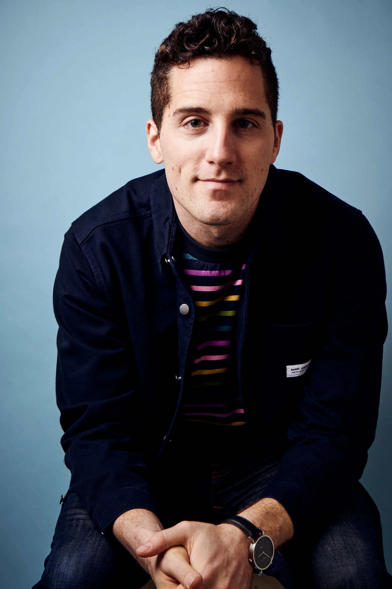 A young man with short curly brown hair and light skin is sitting against a blue background. He is wearing a navy jacket with a label on the chest, a multicolored striped shirt underneath, and a black wristwatch. He has a slight smile and is looking at the camera.