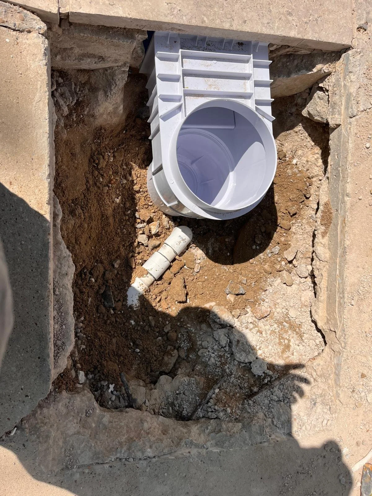 A partially installed white plastic drain pipe in a square hole in the ground surrounded by dirt and small rocks, with a section of a brick wall at the top.