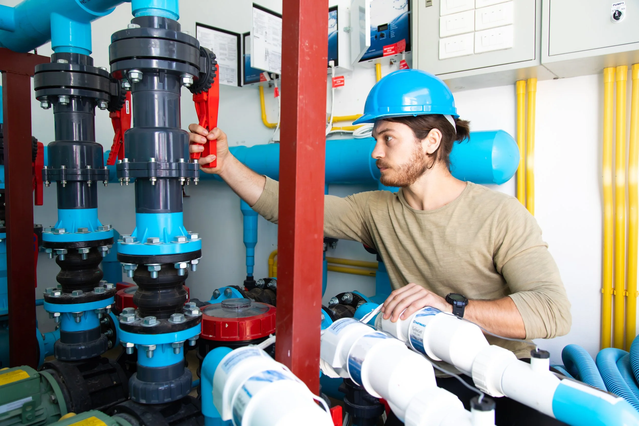A technician wearing a blue hard hat inspects plumbing and piping system with black, blue, and white pipes, valves, and control panels in an industrial setting.