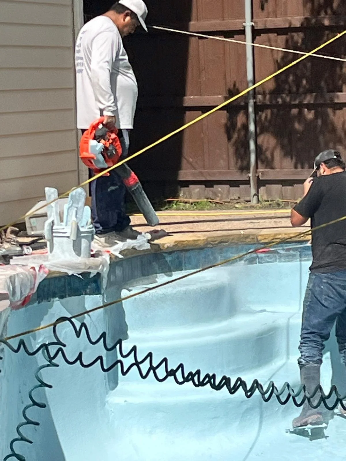 Two men working on the edge of an empty swimming pool, one with a leaf blower and the other with a camera, on a nighttime scene outside a house.