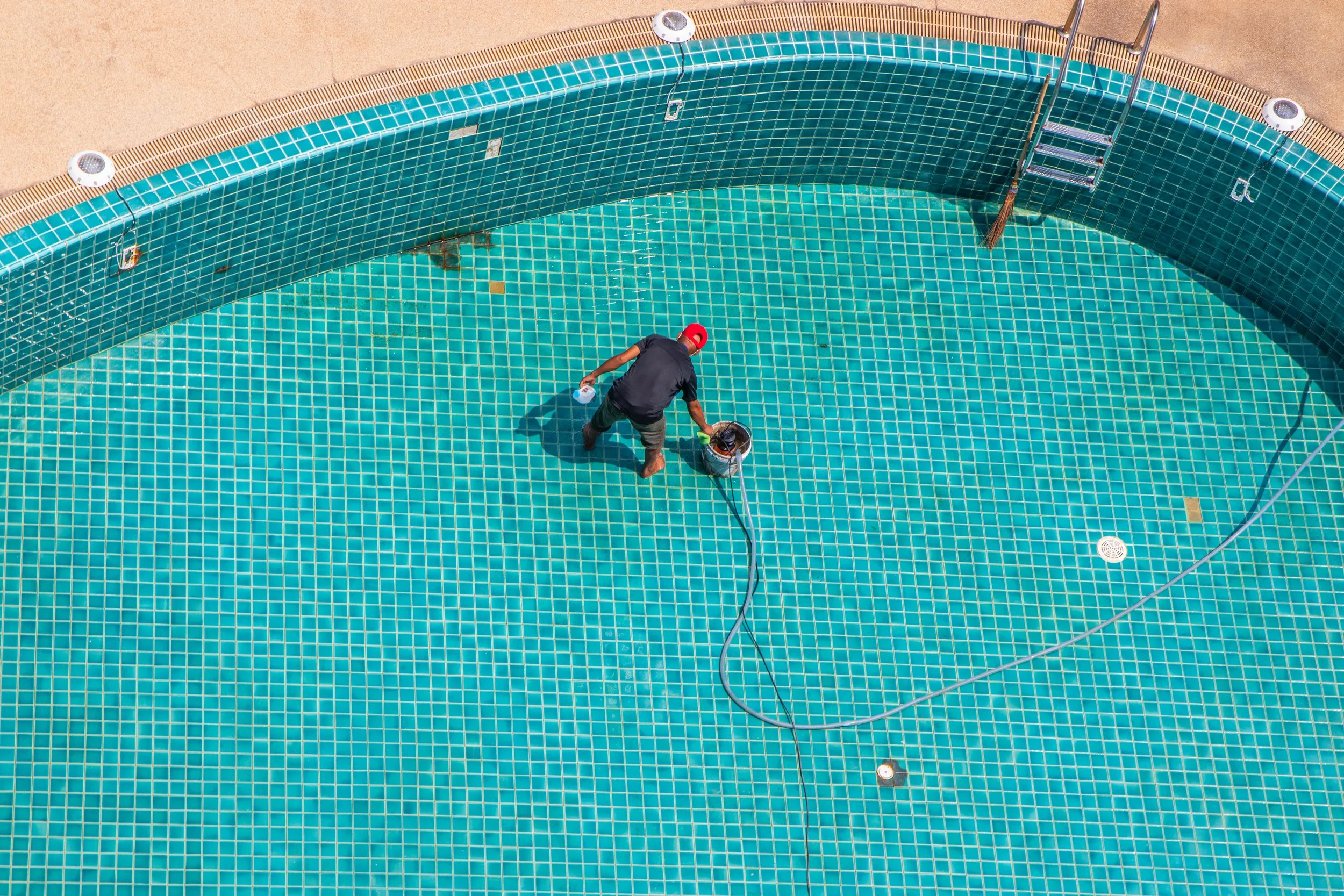 An overhead view of a person cleaning the bottom of a swimming pool with turquoise tiles, a bucket, and a cleaning tool, near a metal staircase.