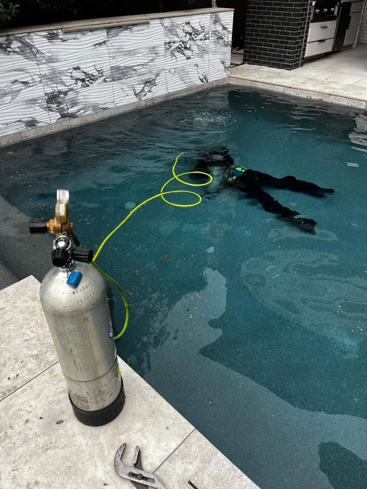 A person in diving gear practicing scuba diving in a swimming pool, with a scuba tank, regulator, and hose visible, along with tools and equipment nearby.
