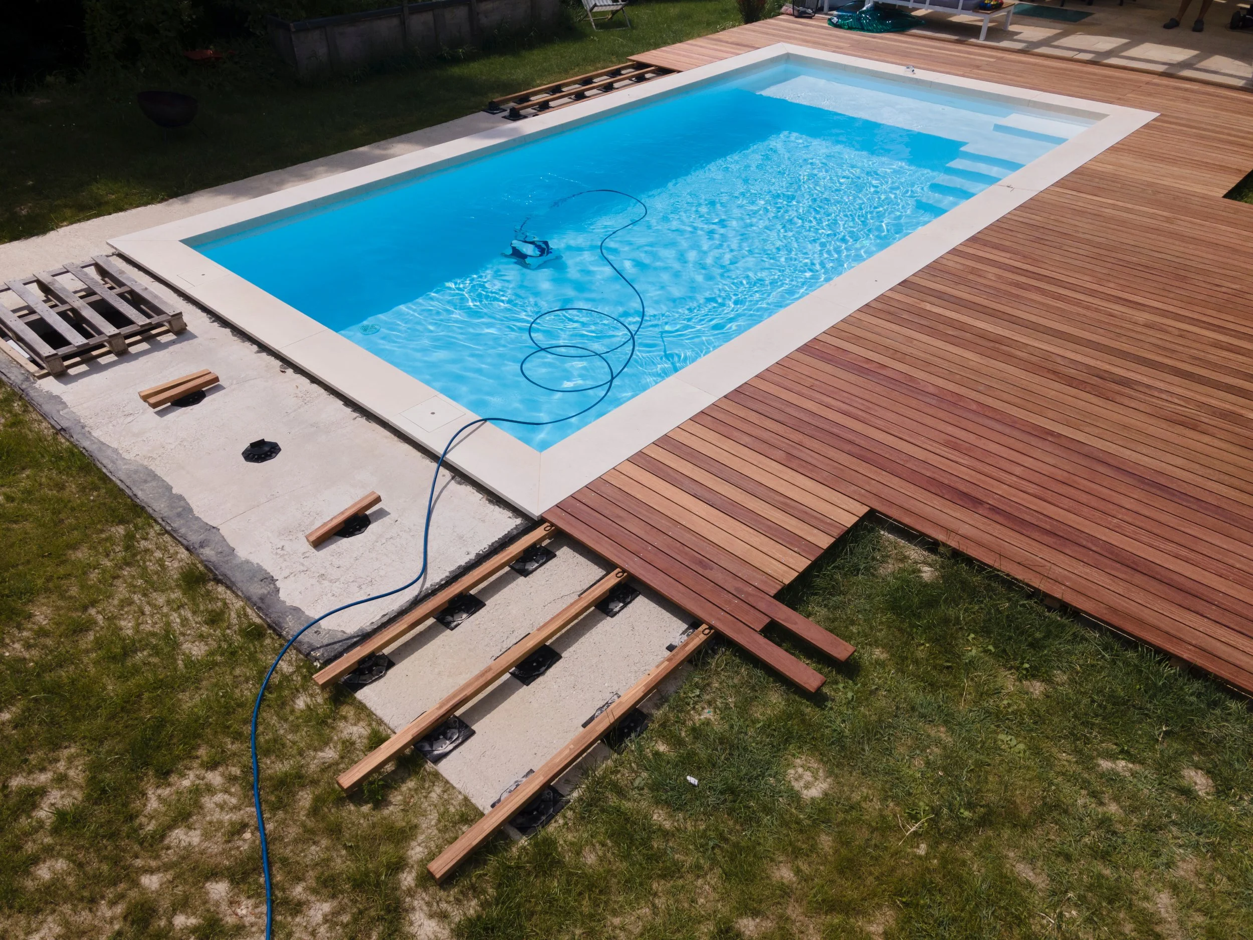 A swimming pool under construction with wooden decking partially installed around it. A robotic pool cleaner is submerged in the pool, and construction materials and tools are visible nearby.