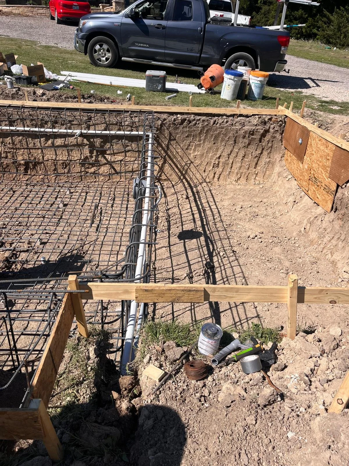 A construction site showing the foundation of a building with exposed rebar and wooden framing. There are tools, pipes, and construction materials scattered around. In the background, there is a gravel driveway with parked vehicles, including a black truck and a red car.