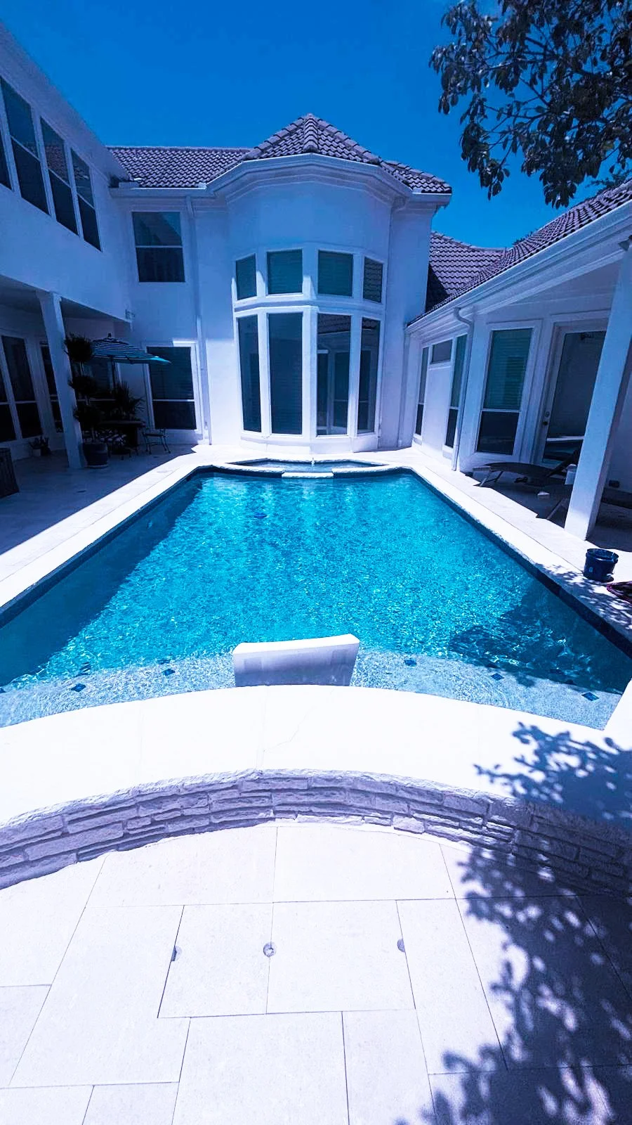 Backyard swimming pool with a white house exterior, large windows, and a patio area during daytime.