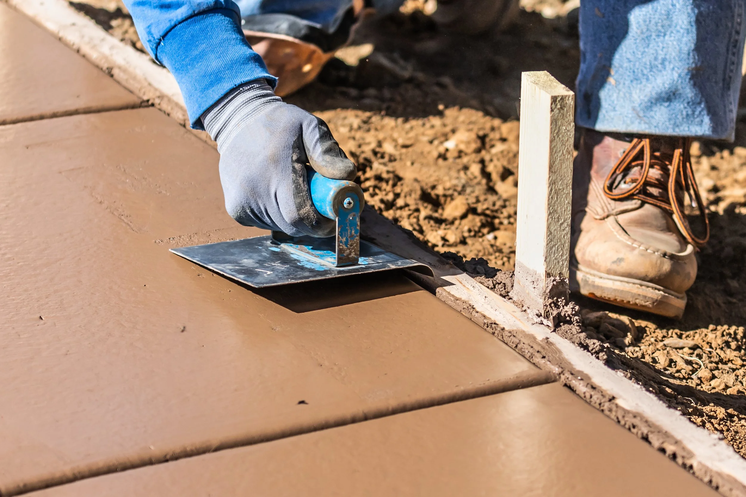 A person wearing work gloves, blue jacket, jeans, and work boots smoothing a new concrete sidewalk with a hand trowel.