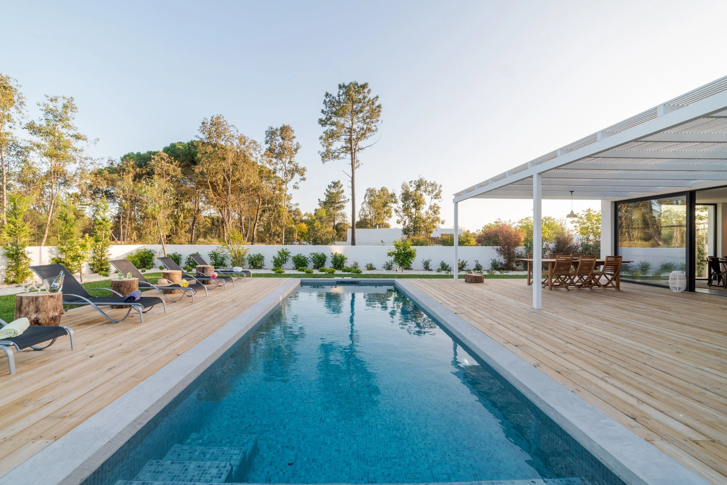 Modern backyard with a rectangular pool, wooden deck, lounge chairs, and outdoor dining area under a white pergola.